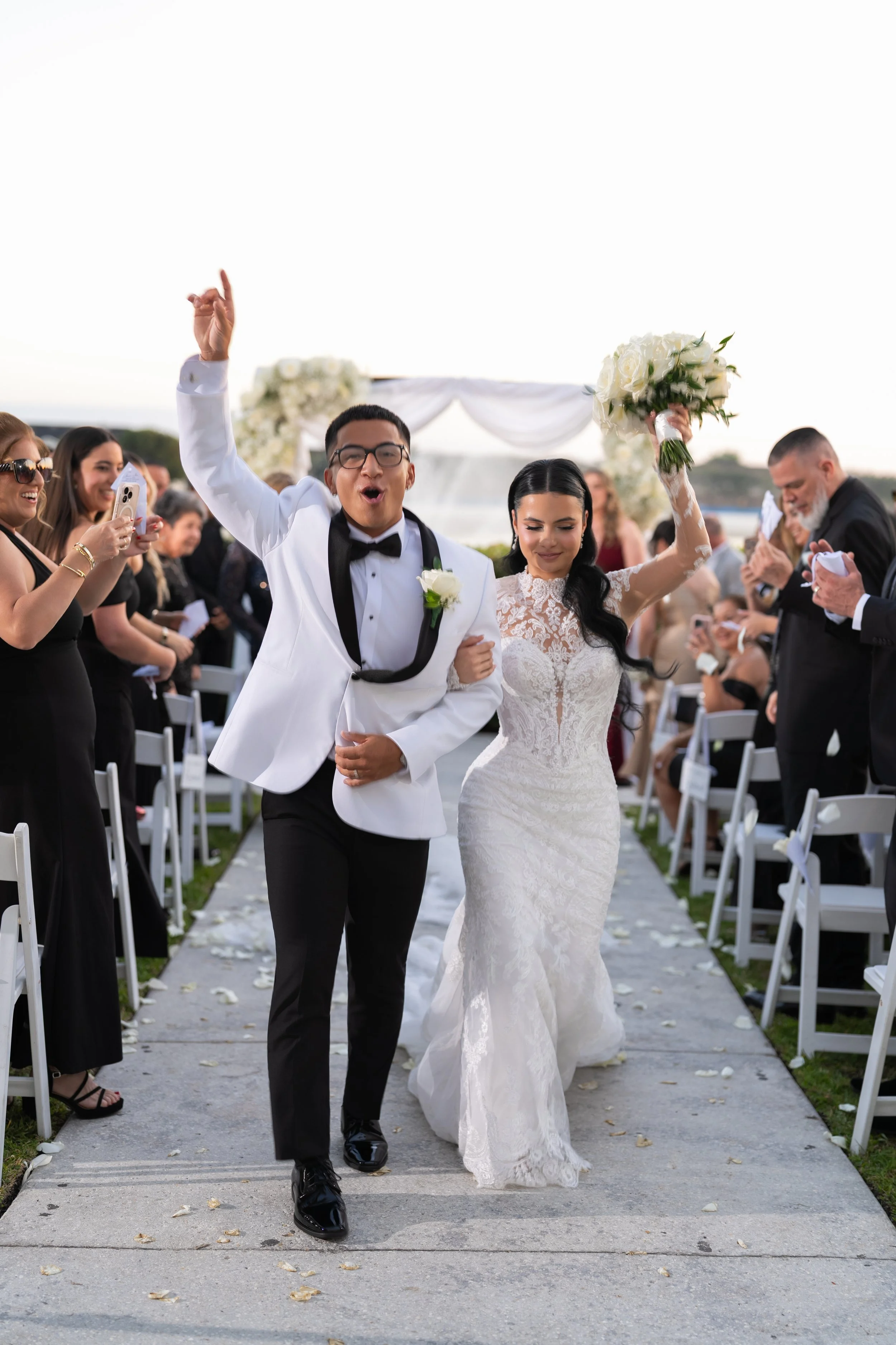A newly married couple walking down the aisle at an outdoor wedding, celebrating with guests, with the bride holding a bouquet and the groom in a tuxedo.