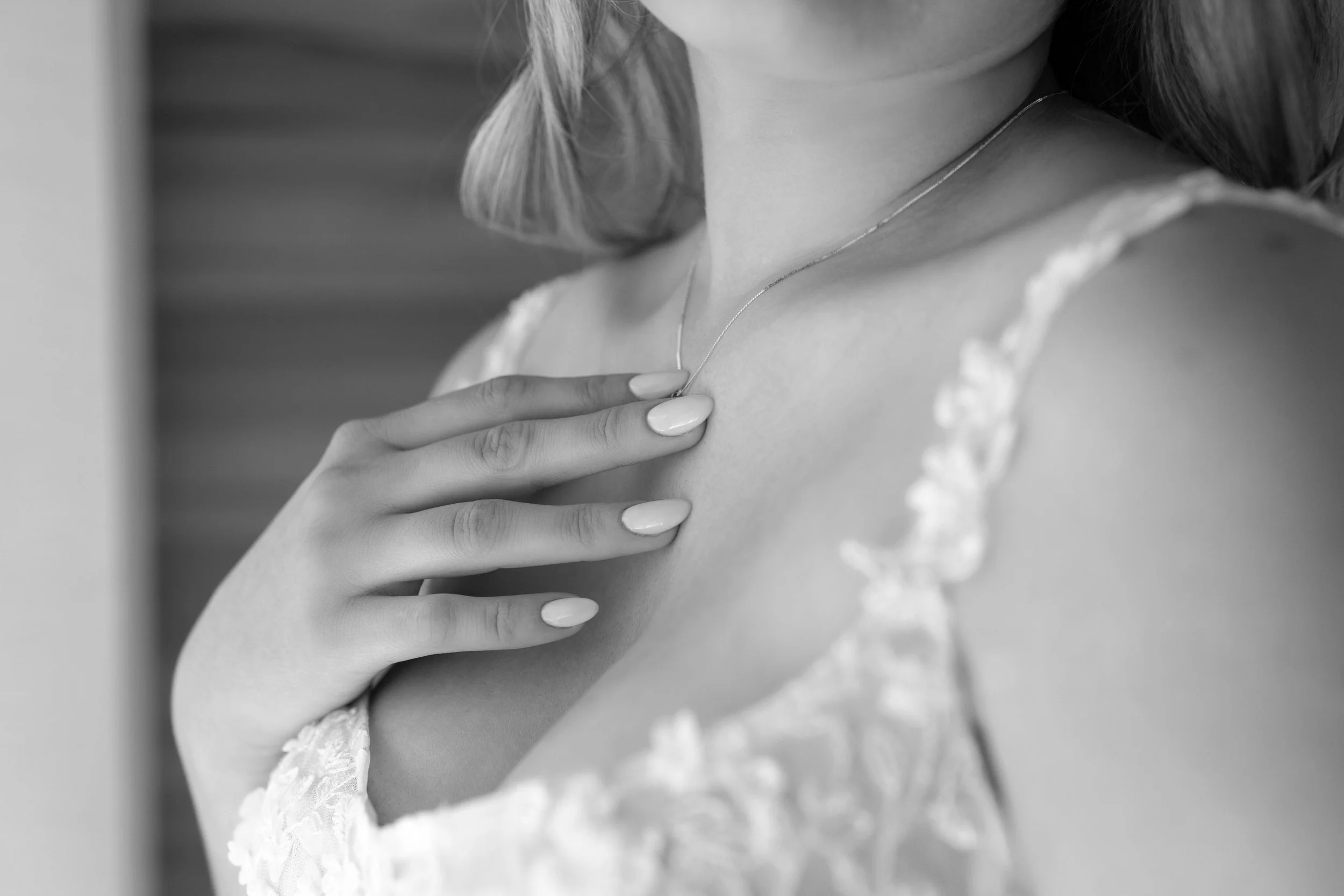 Close-up of a woman wearing a lace dress and a delicate necklace, with her hand placed on her chest, in black and white.