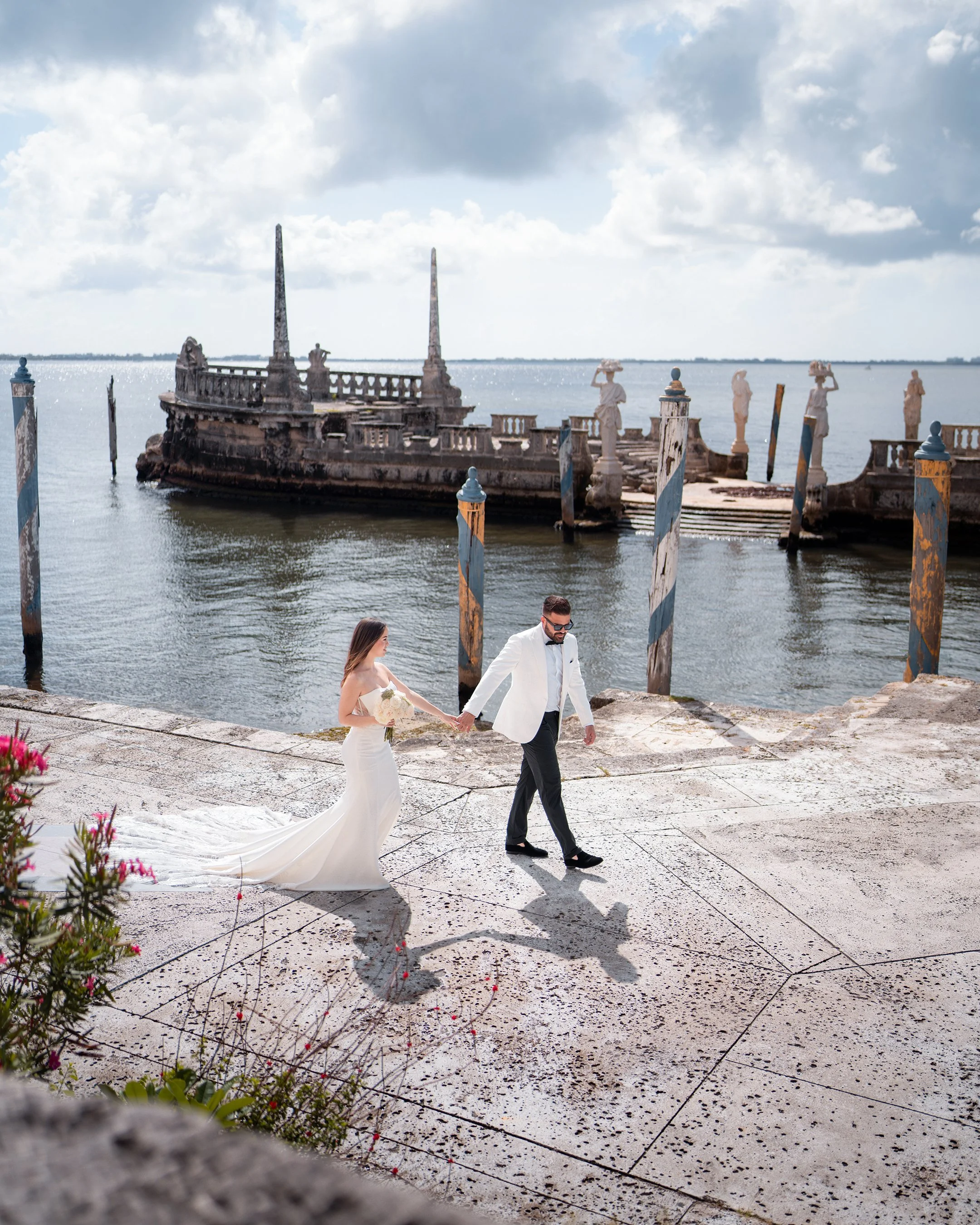 Bride and groom walking hand in hand along a waterfront with an old, weathered boat docked by the pier in the background.