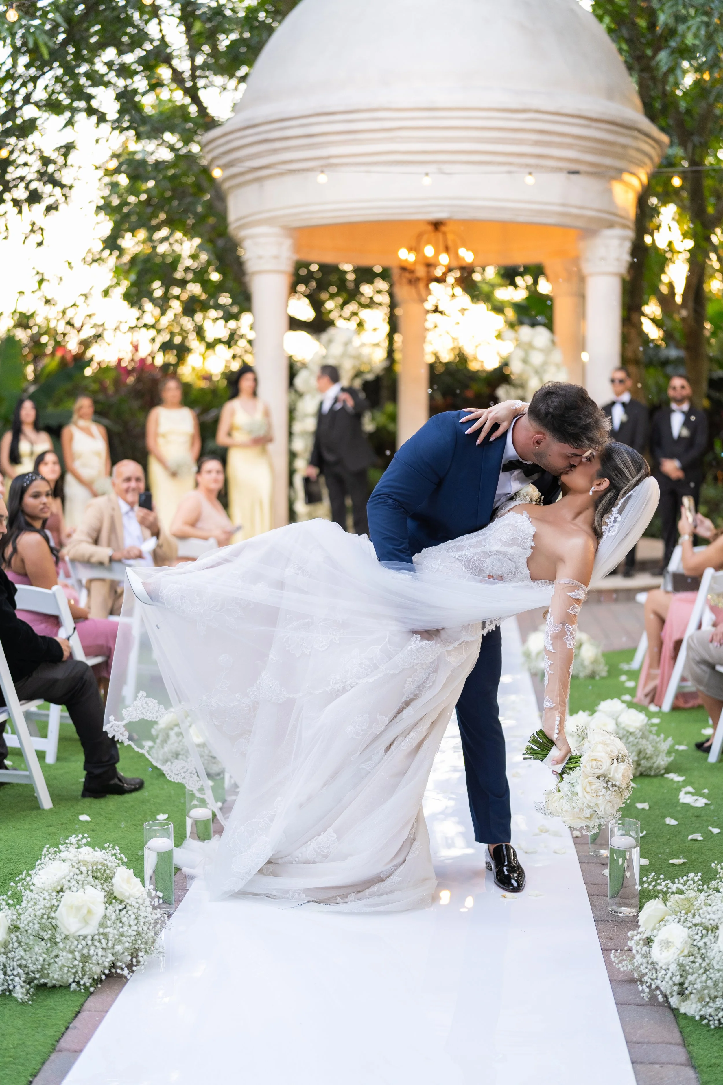A bride and groom share a kiss during their outdoor wedding ceremony, with guests seated on either side and a white gazebo in the background.