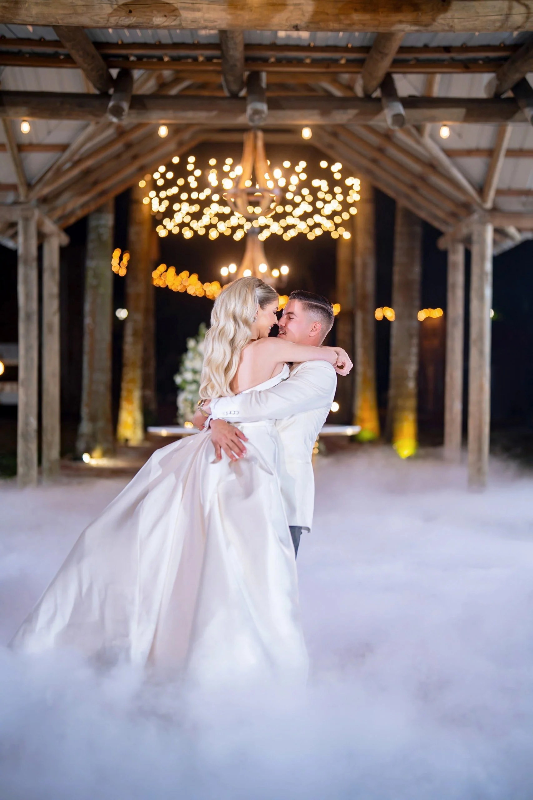 A bride and groom dance closely under string lights and a chandelier at a rustic barn wedding, with fog on the ground.