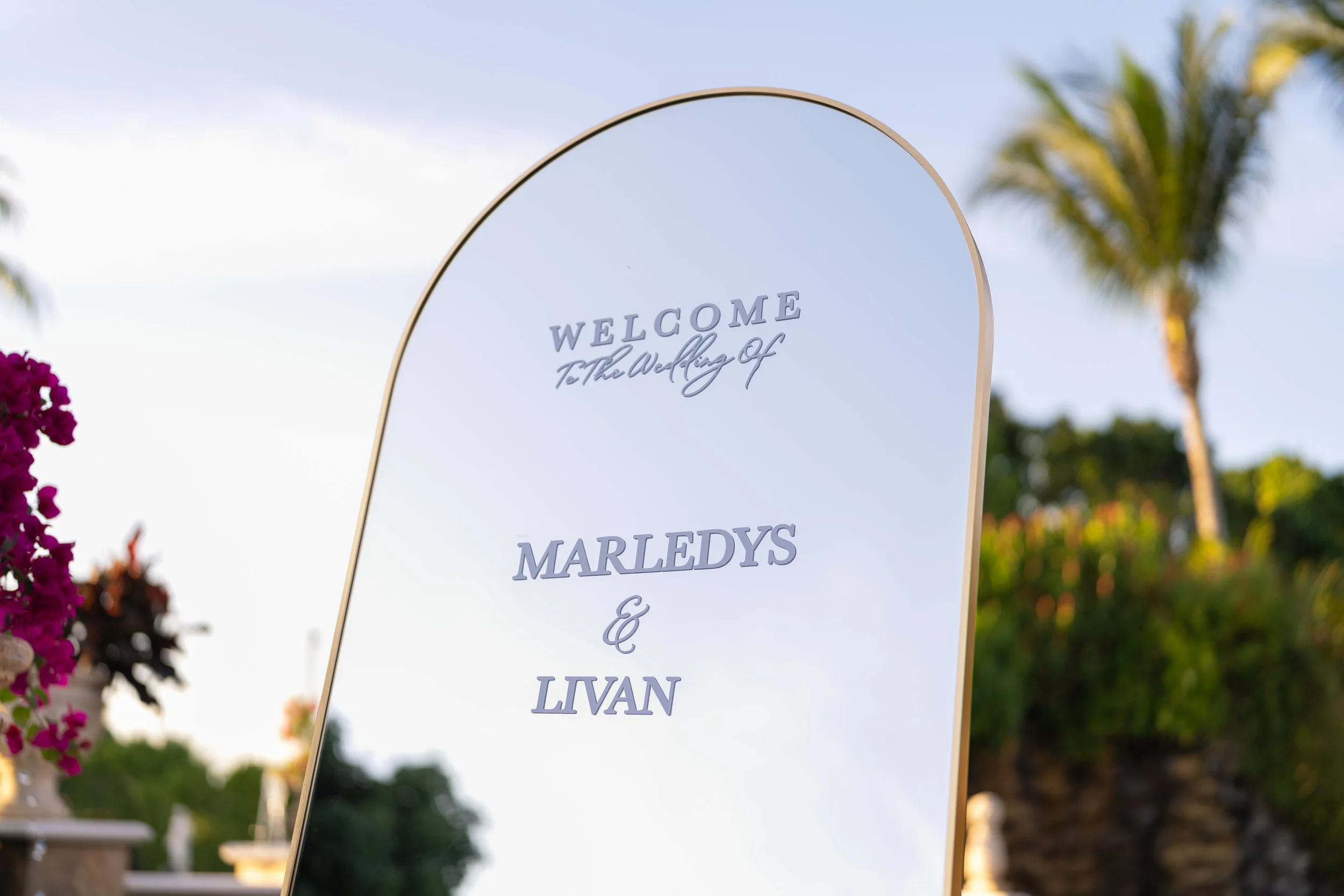 Wedding welcome sign on a reflective mirror with trees and flowers in the background.