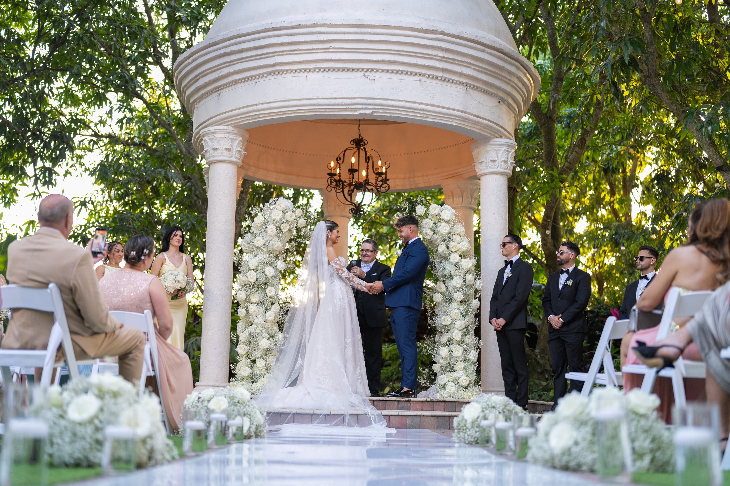 A wedding ceremony taking place outdoors under a small white pavilion decorated with white flowers and greenery. The bride and groom are holding hands and smiling at each other, standing in front of a officiant. Guests are seated around, some taking 