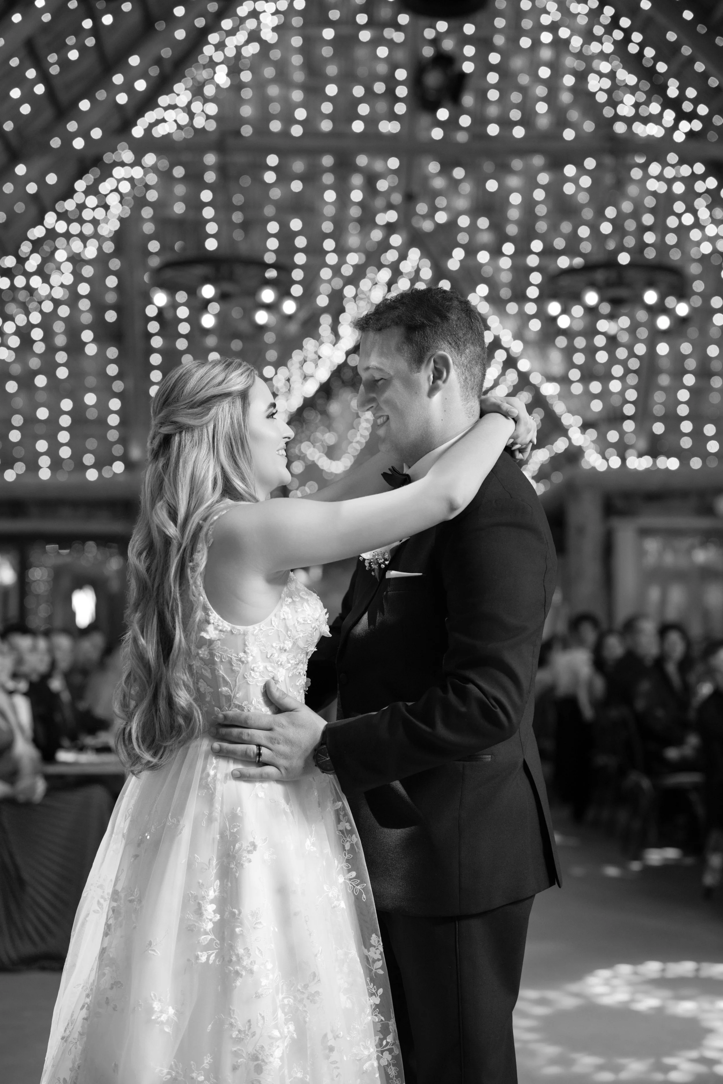 A bride and groom dancing at their wedding reception under string lights, with guests seated in the background.