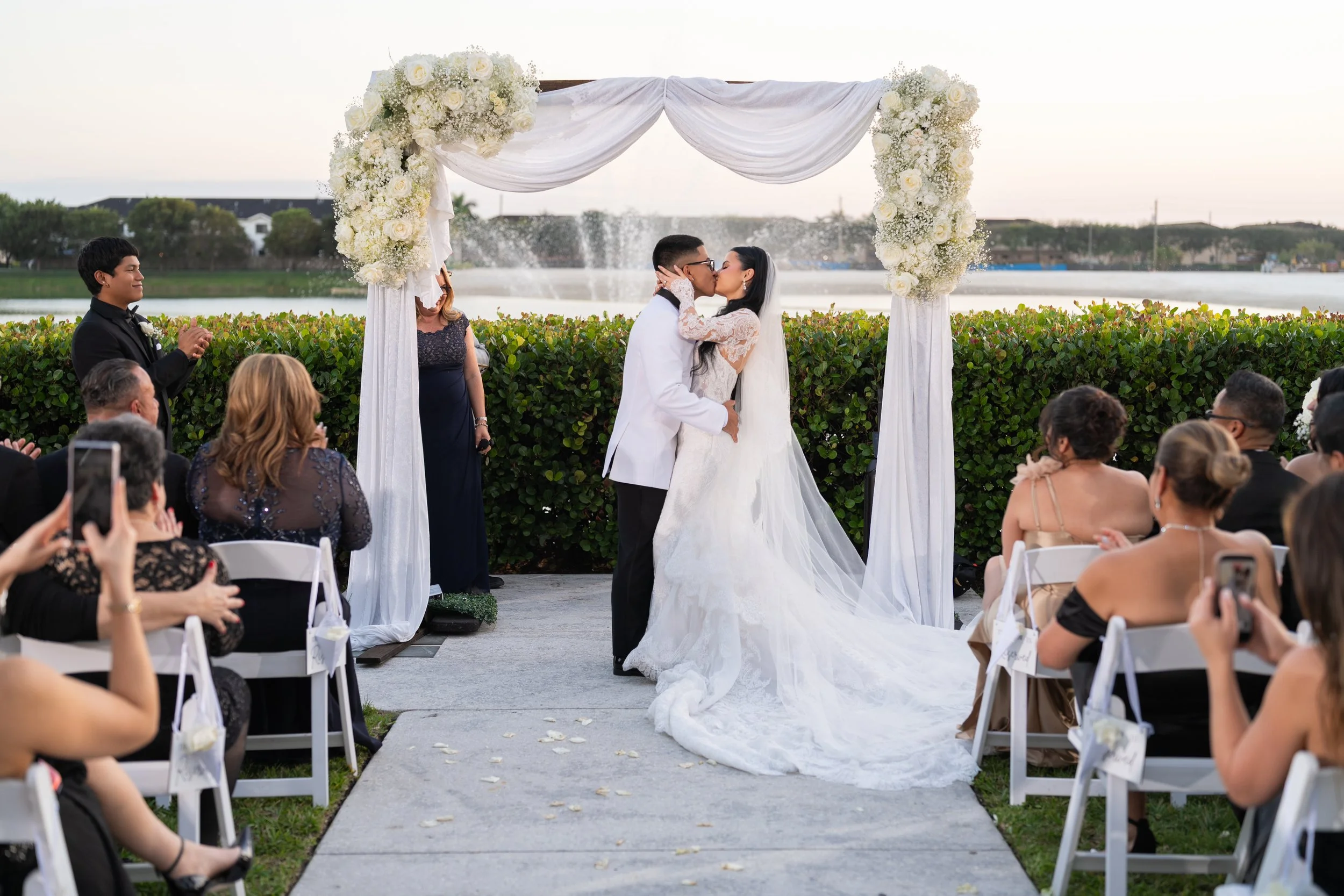A wedding ceremony outdoors with a couple kissing under a white floral arch as guests watch and take photos by a lake in the background.