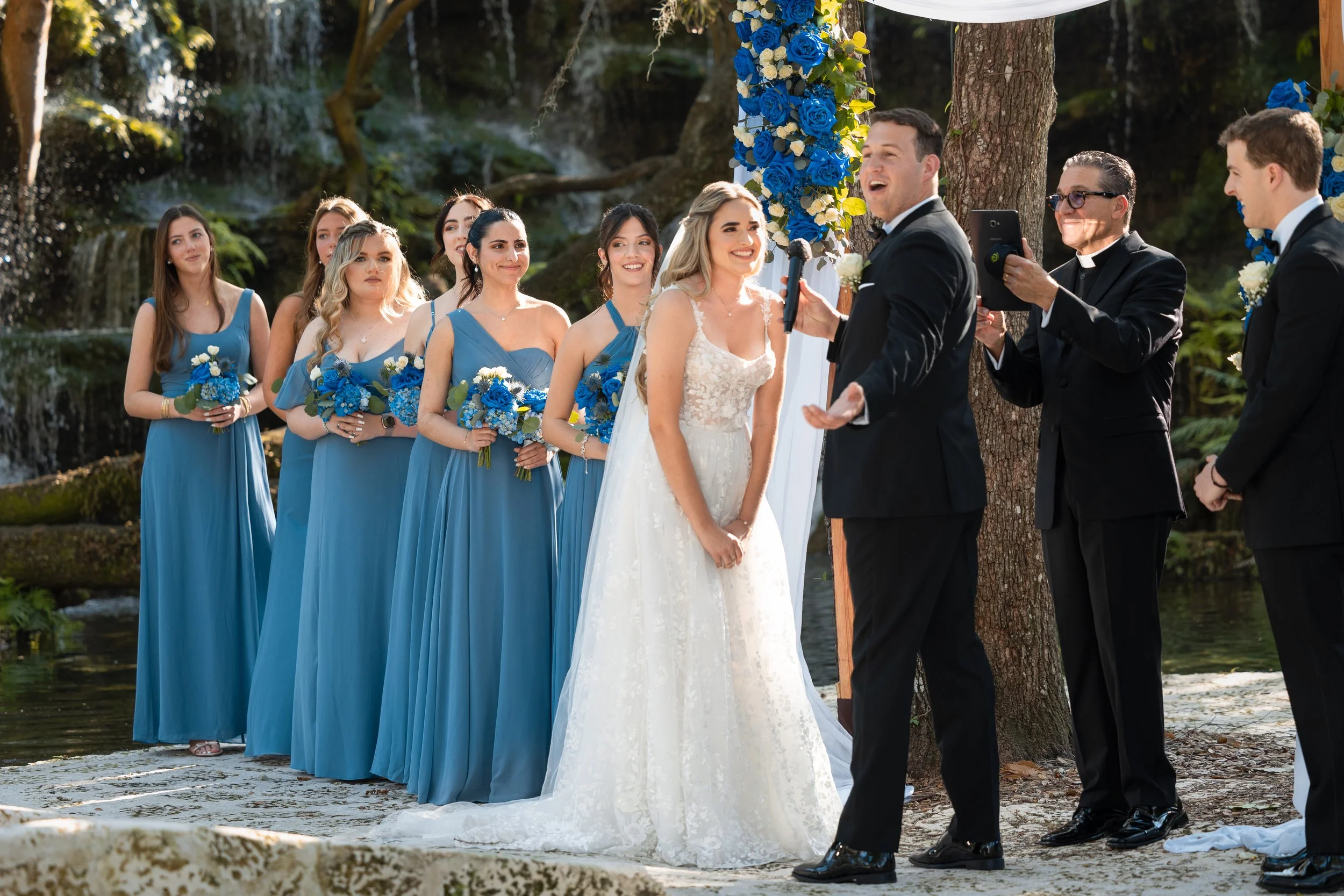 A wedding ceremony taking place outdoors near a waterfall, with the bride and groom facing each other, surrounded by bridesmaids in matching blue dresses, officiant, and guests.