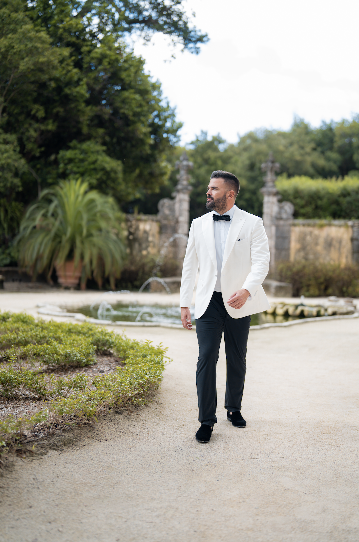 A man in a white tuxedo jacket and black pants walking outside near a fountain and greenery.