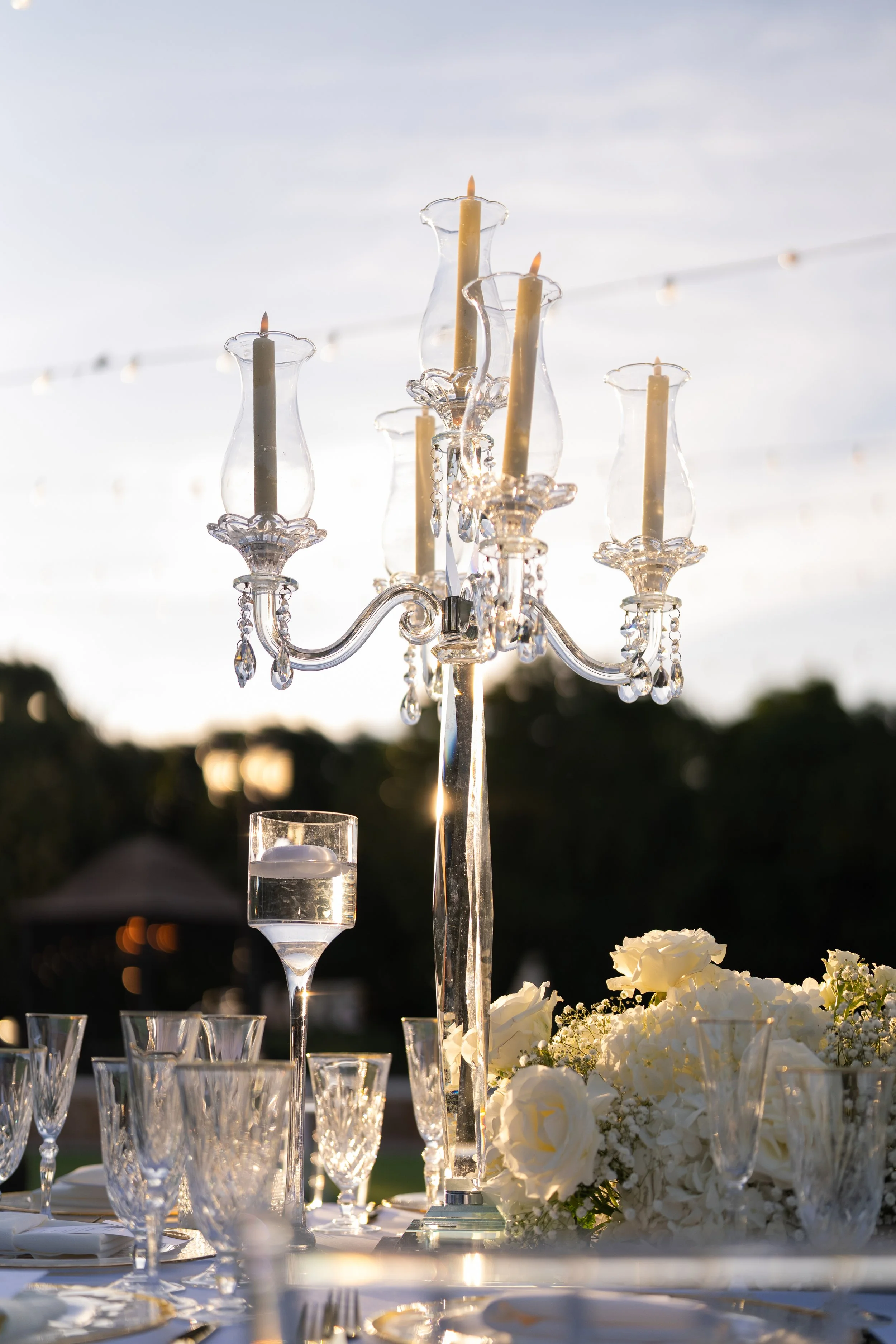 Elegant outdoor table setting with a crystal chandelier centerpiece, surrounded by white flowers, glassware, and a sunset background.