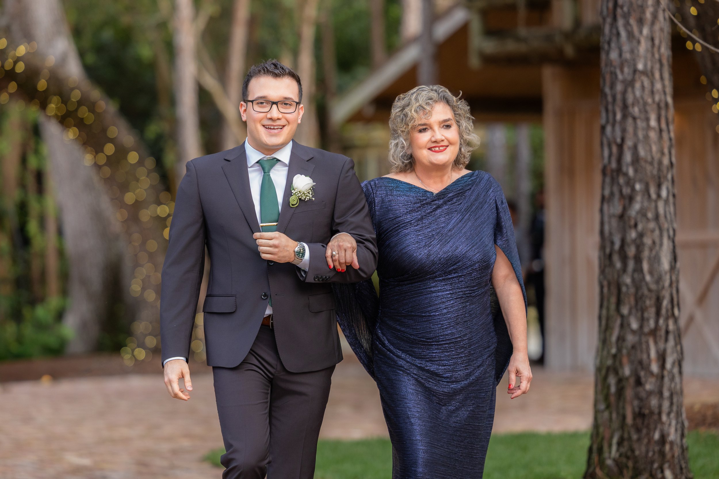 A young man in a black suit, white shirt, and green tie walking arm-in-arm with a woman in a dark blue dress outdoors at a formal event or wedding, with string lights and trees in the background.
