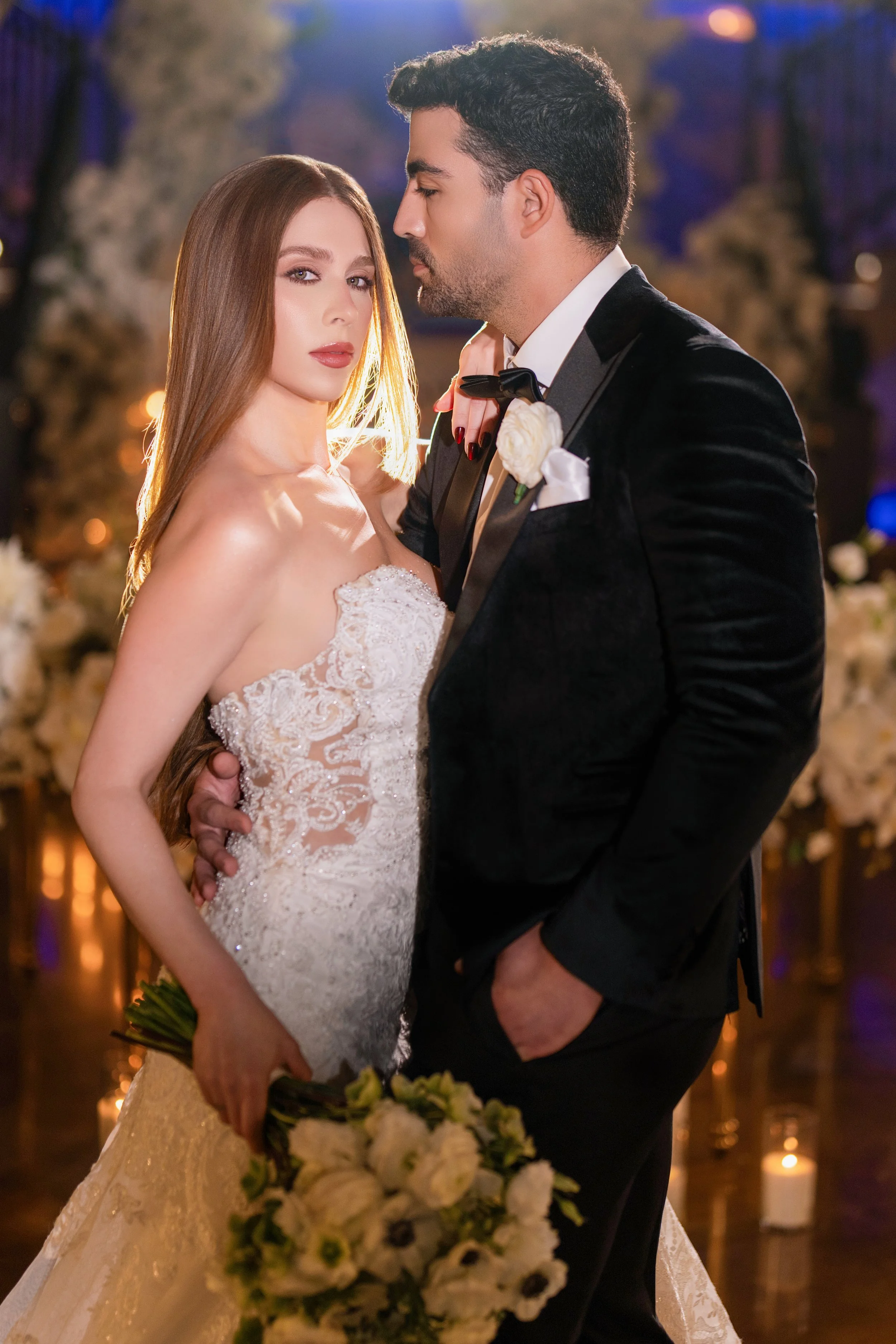 A bride and groom standing close together at their wedding, with the bride holding a bouquet of flowers and the groom in a tuxedo, in a decorated indoor venue.