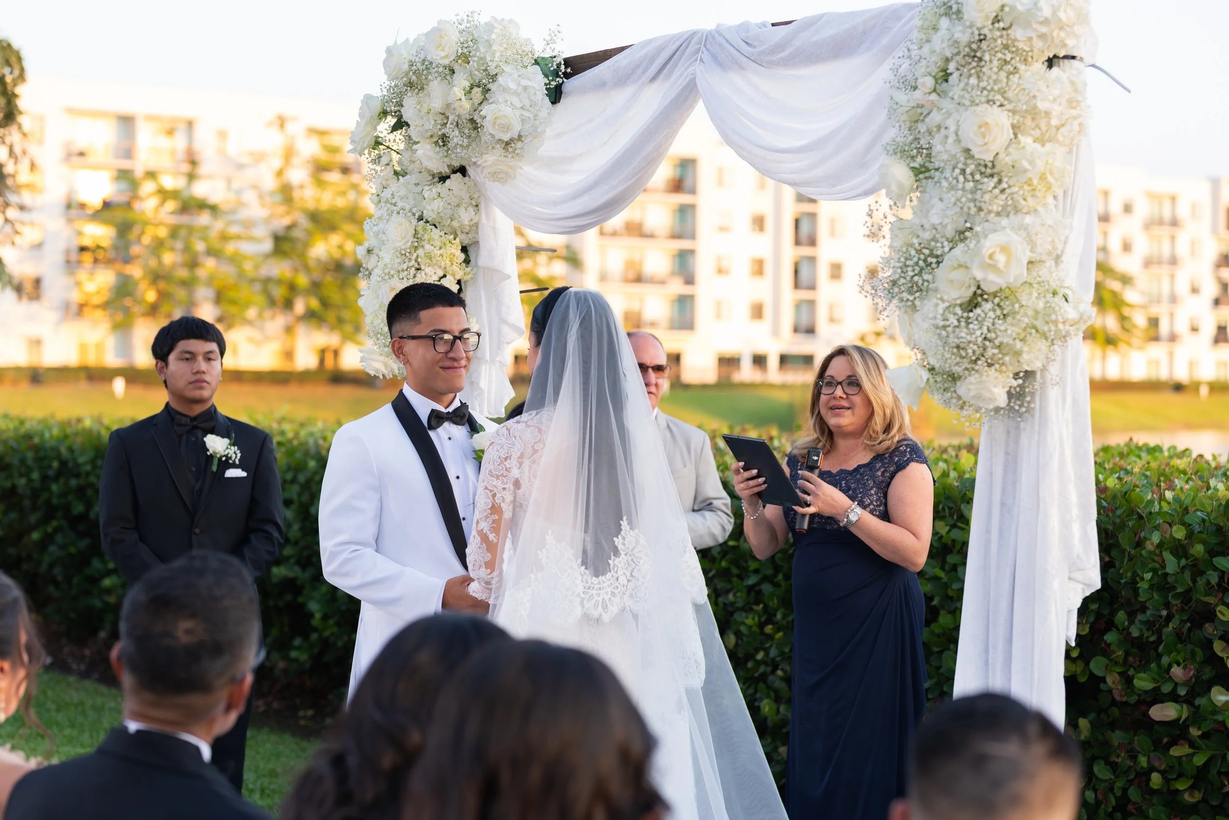 A wedding ceremony outdoors with a couple exchanging vows under a floral arch, surrounded by guests.