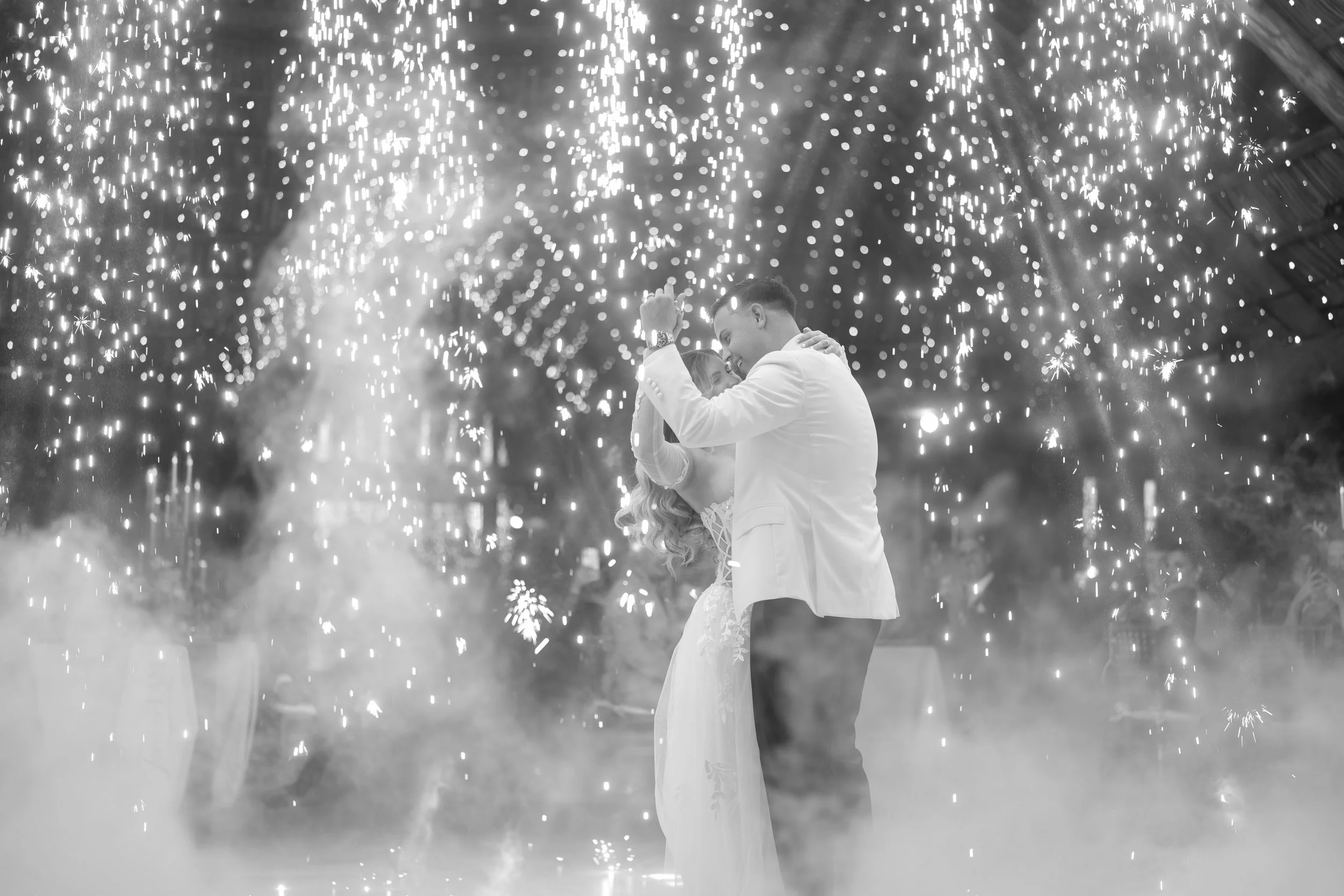 A couple at their wedding dance surrounded by sparklers and smoke, in a festive celebration setting.