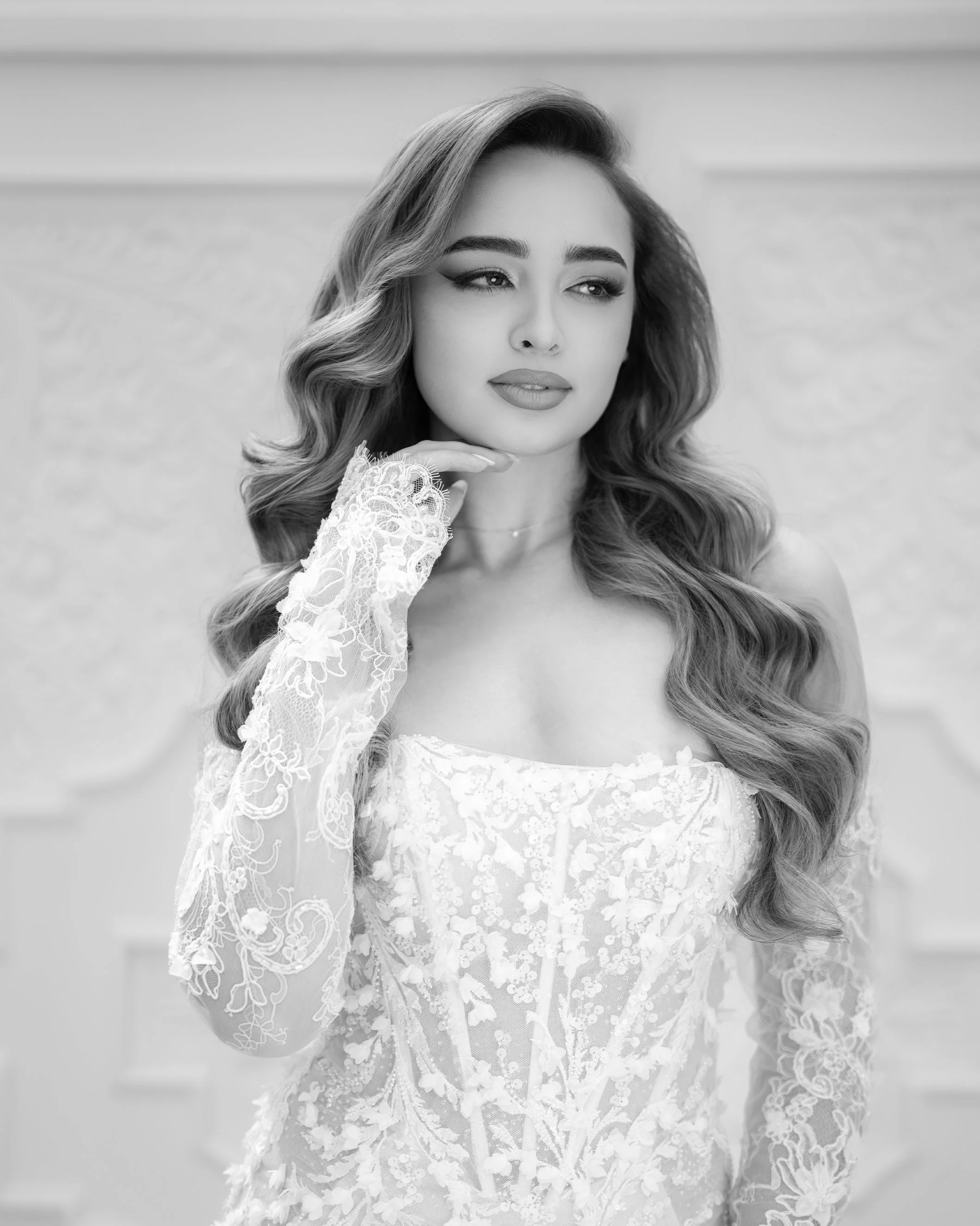 Black and white portrait of a woman in a lace wedding dress with bridal makeup and wavy hair, posing indoors.