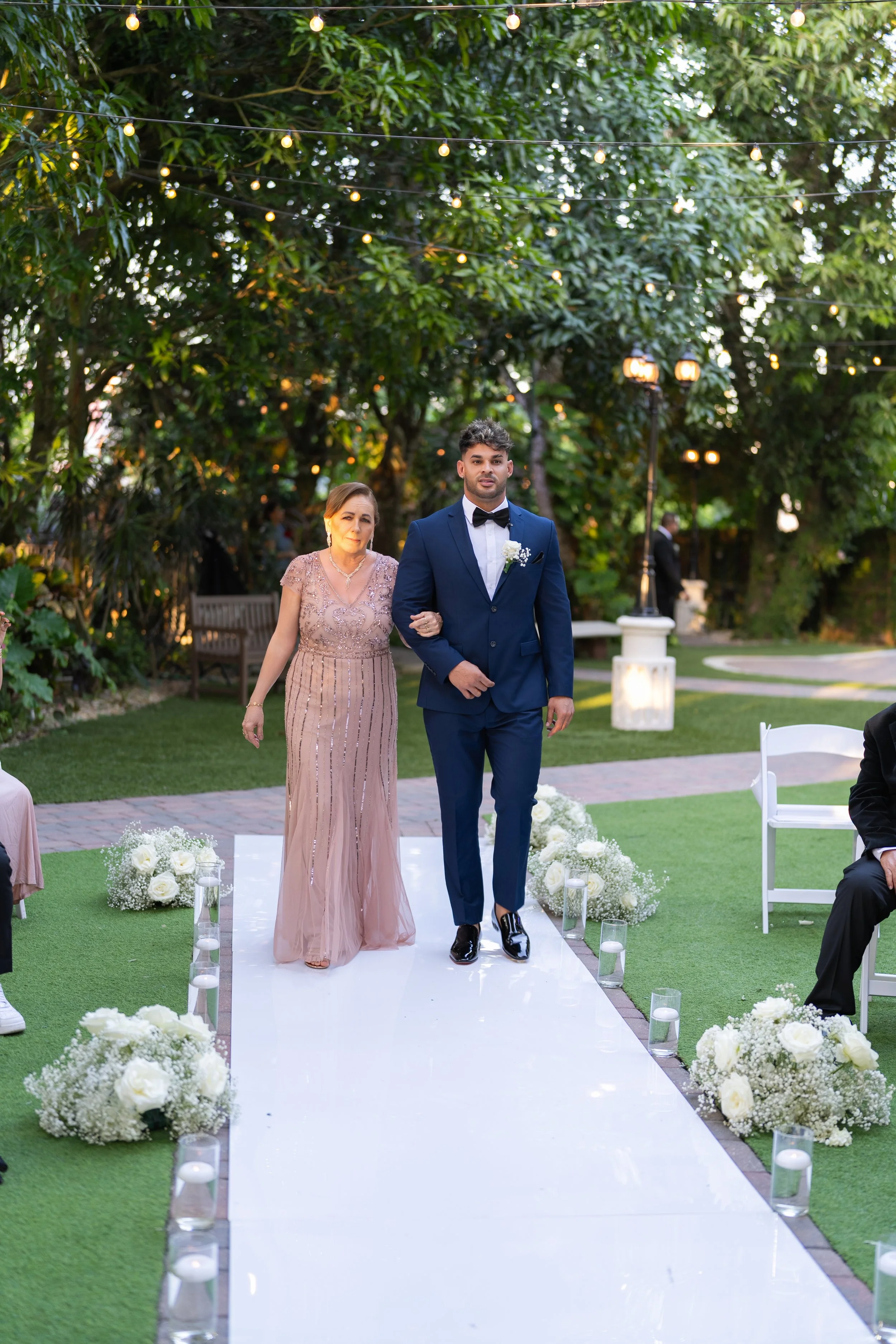 A young man in a navy blue suit and bow tie walks down an outdoor wedding aisle accompanied by an older woman in a pink gown. The aisle is decorated with white flower arrangements and candles, set in a lush garden with string lights overhead.