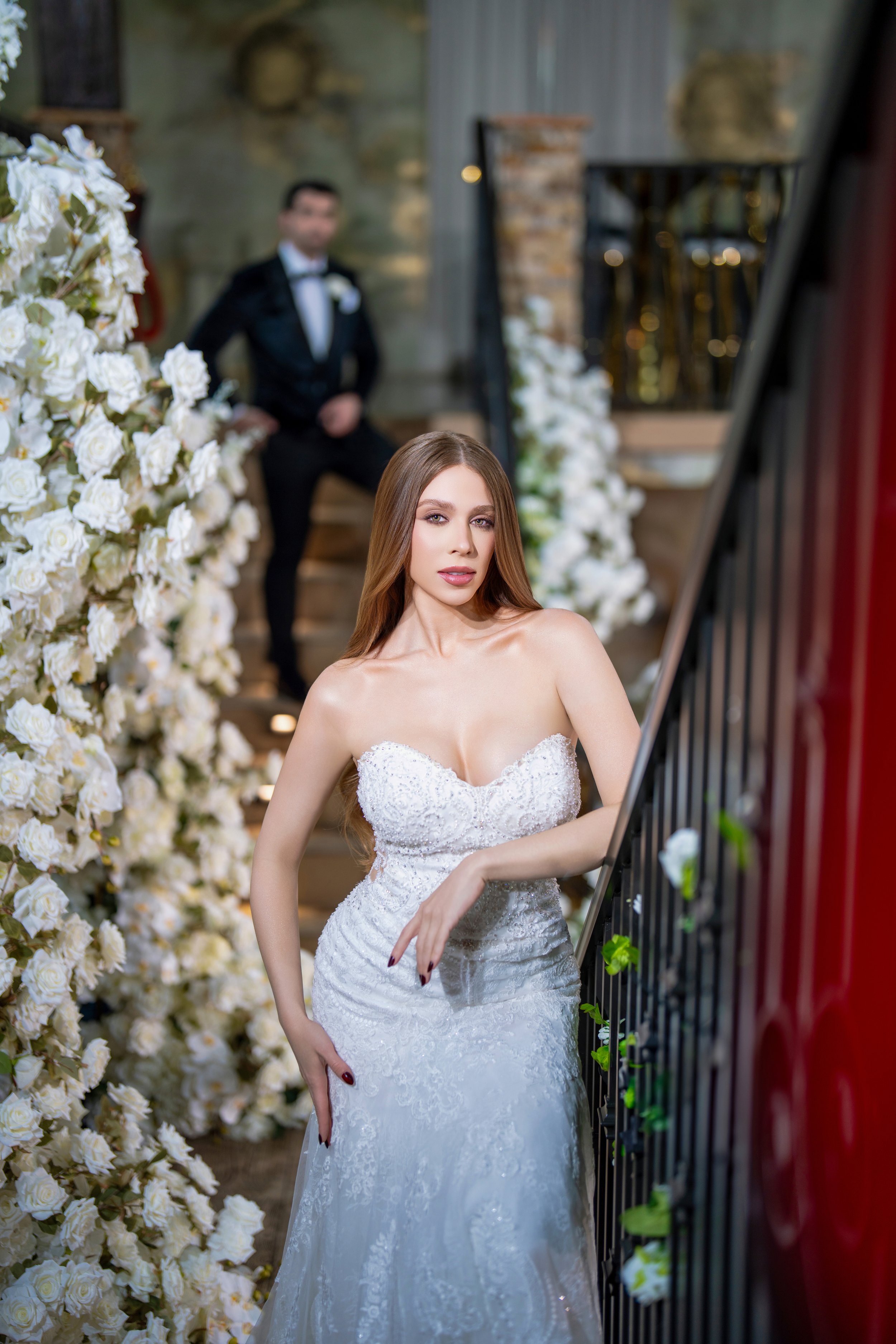 A woman in a strapless wedding dress standing on a staircase, with a man in a tuxedo sitting on the stairs behind her, surrounded by white flowers.