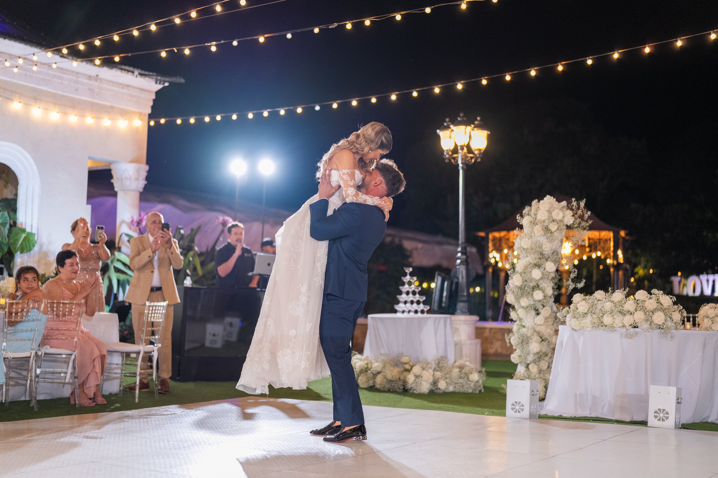 A bride and groom share a dance at their wedding reception outdoors at night, surrounded by guests taking photos and colorful string lights.