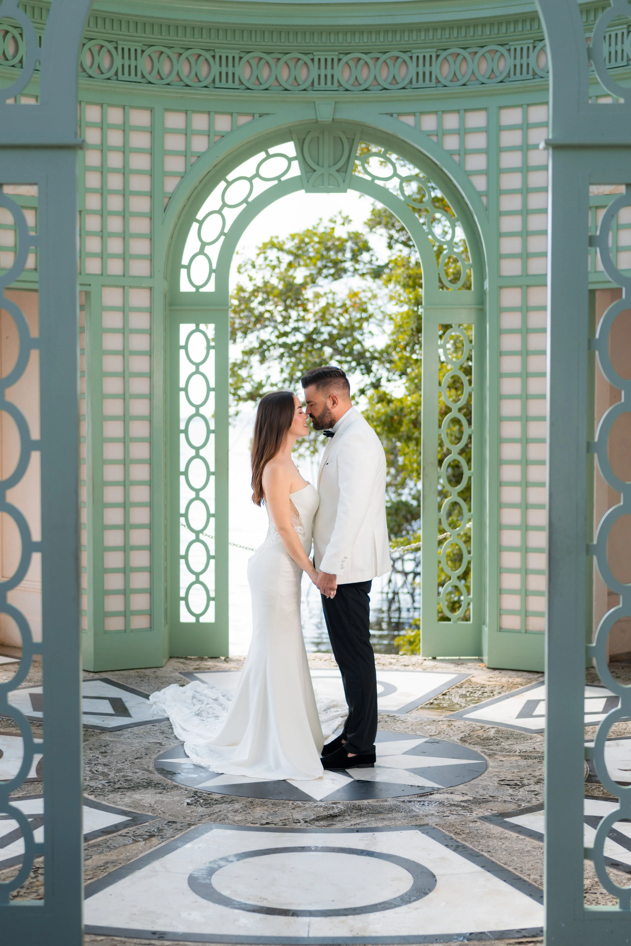 A bride and groom holding hands and leaning into each other standing under a decorative arch at an outdoor wedding venue. The bride is wearing a strapless white wedding gown and the groom is in a white tuxedo jacket with black pants, both are looking