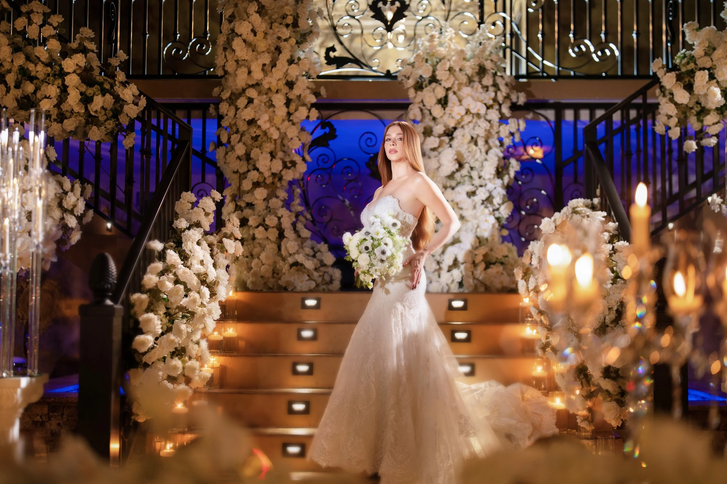 A bride in a white wedding gown holding a bouquet of white flowers stands on a grand staircase decorated with white flowers and candles, surrounded by floral arrangements and ornate black railings.
