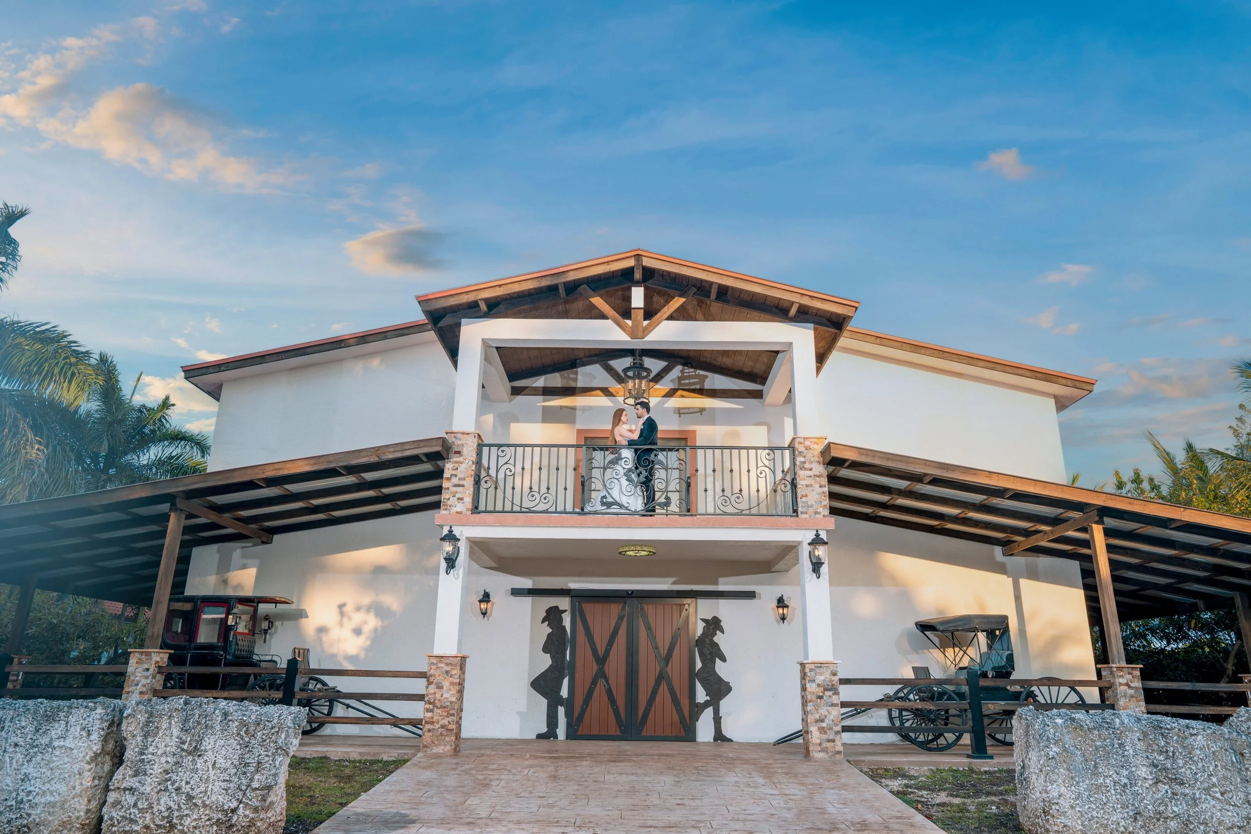 A couple standing on a balcony of a large, white, two-story house with rustic accents, holding hands and facing each other during sunset with a cloudy sky, trees, and vintage decor including a wagon and lanterns.