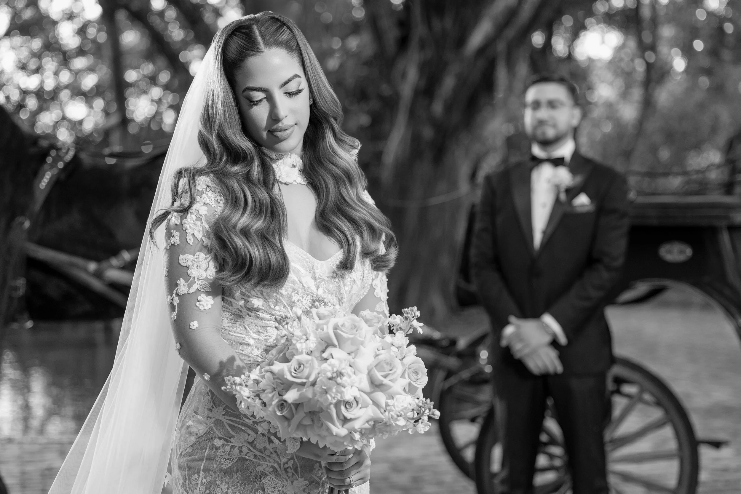A bride holding a bouquet of roses and flowers, with a groom standing behind a vintage horse-drawn carriage in a park.