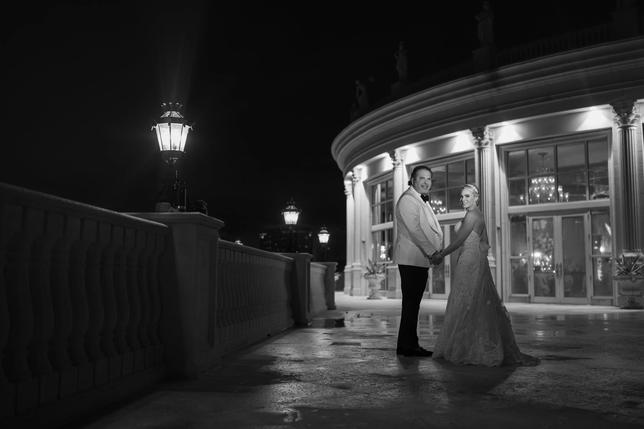 A couple in wedding attire holding hands and smiling at each other outside a lit building at night.