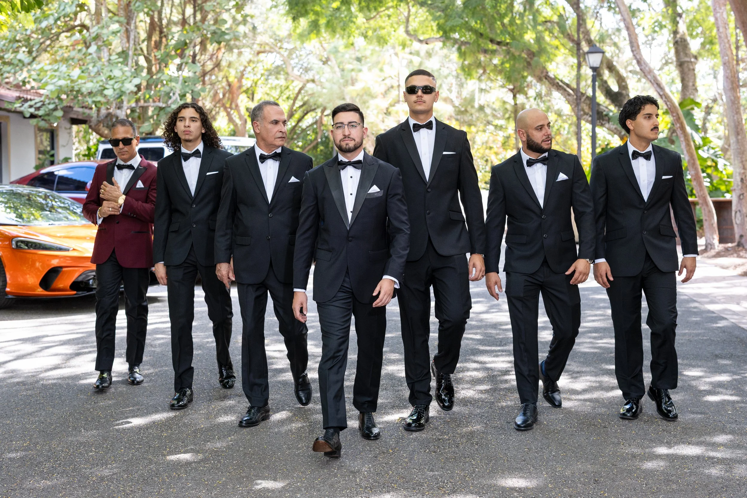 Group of men dressed in formal tuxedos walking outdoors, with a luxury sports car in the background, trees, and a lamppost.