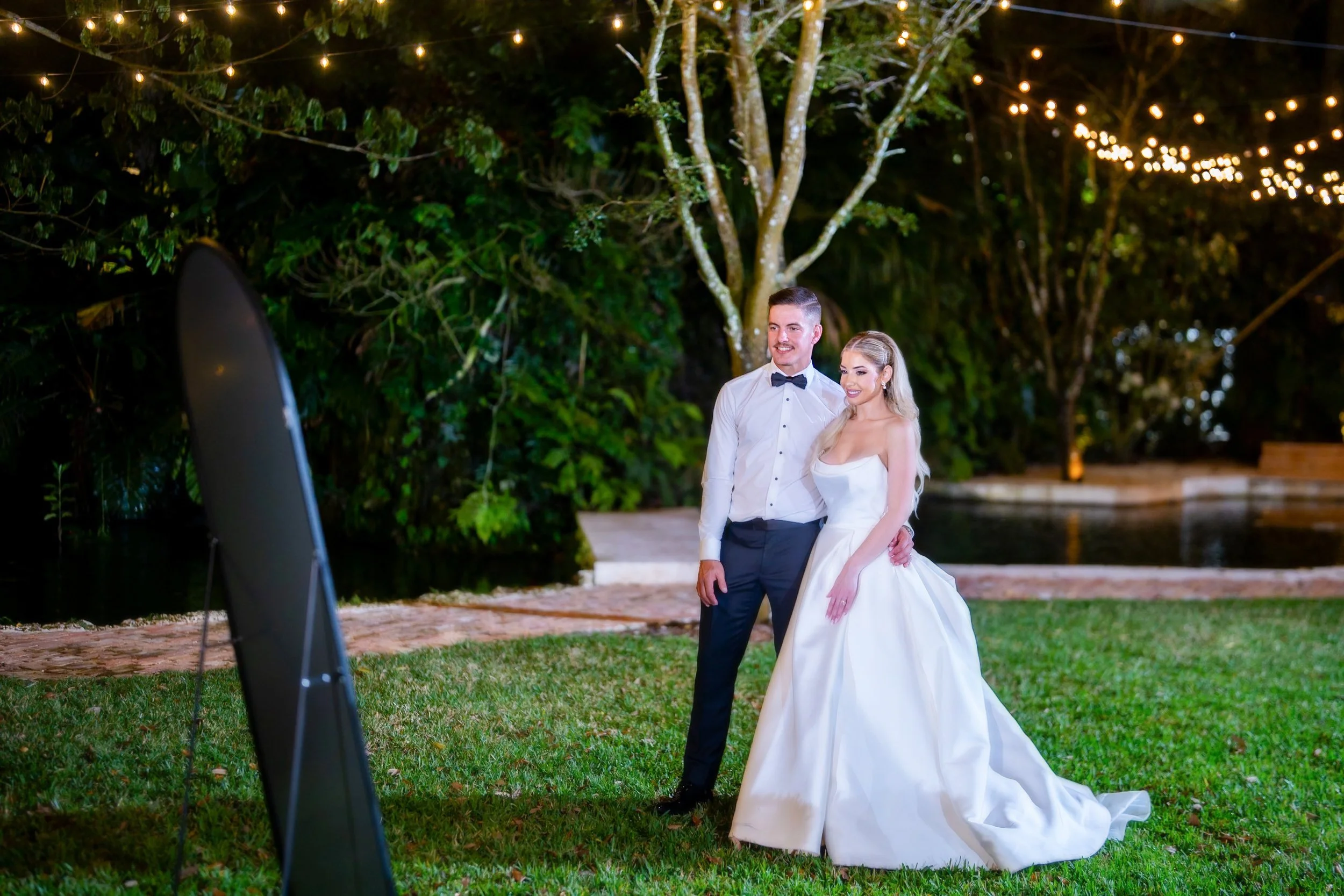 A newlywed couple taking their wedding photos outdoors at night. The bride is in a white wedding gown, and the groom is in a white shirt with a black bow tie and dark pants. They are standing on grass near a pond, with trees and string lights in the 