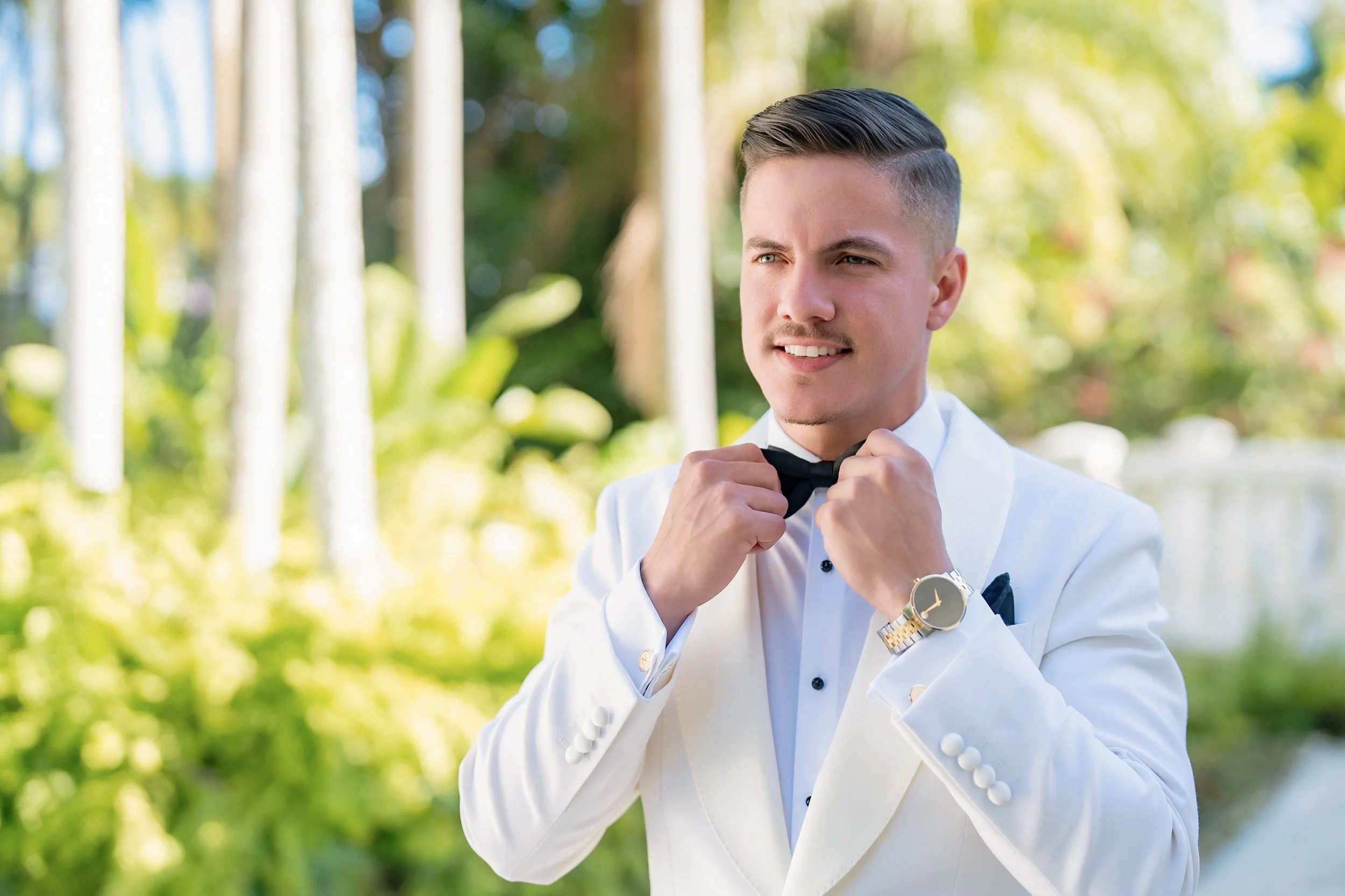 A young man in a white tuxedo adjusting a black bow tie outdoors with lush green trees and sunlight in the background.