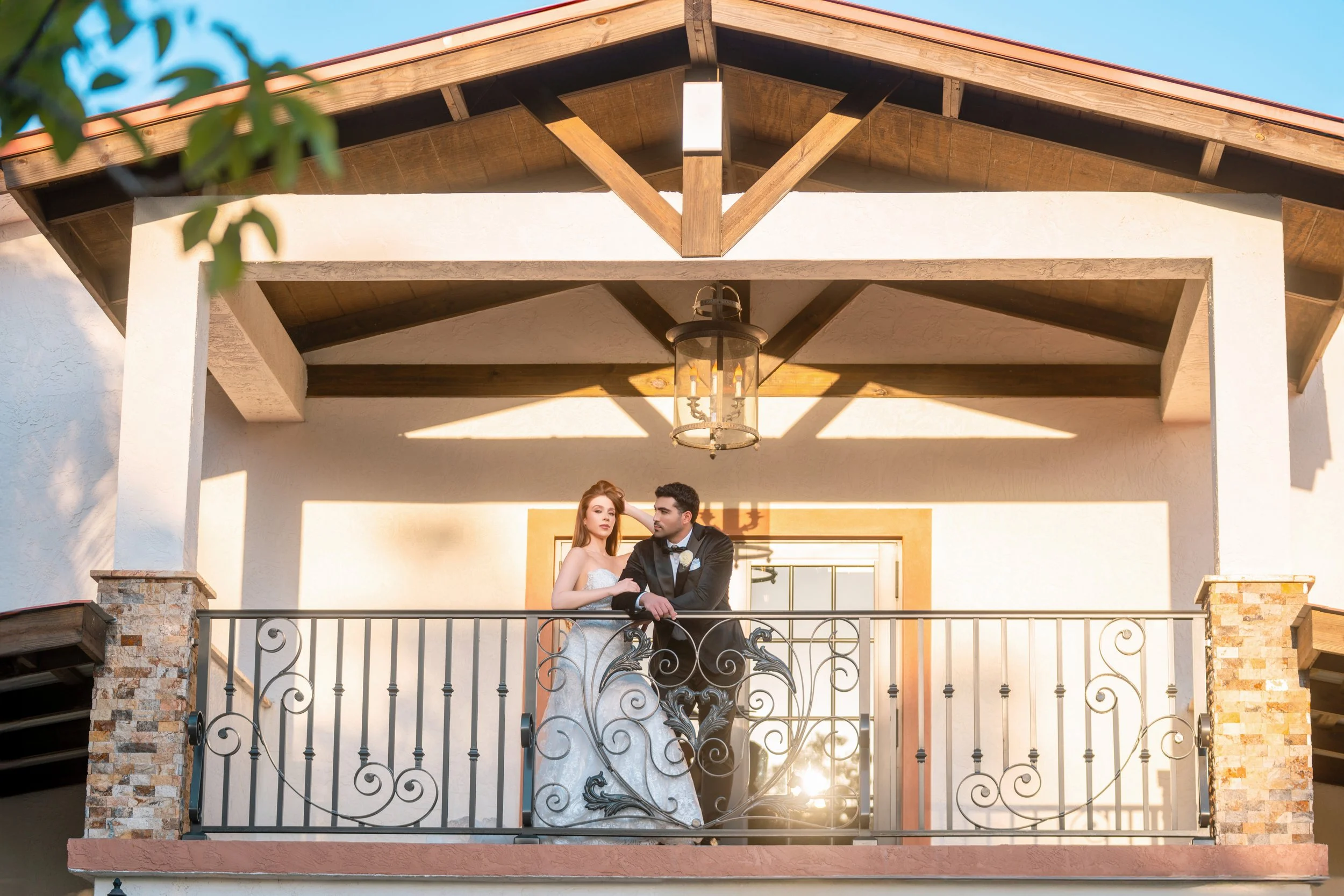A bride and groom standing on a balcony with intricate black wrought iron railing, under a wooden and stone sheltered patio, with sunlight casting shadows on the white wall behind them.