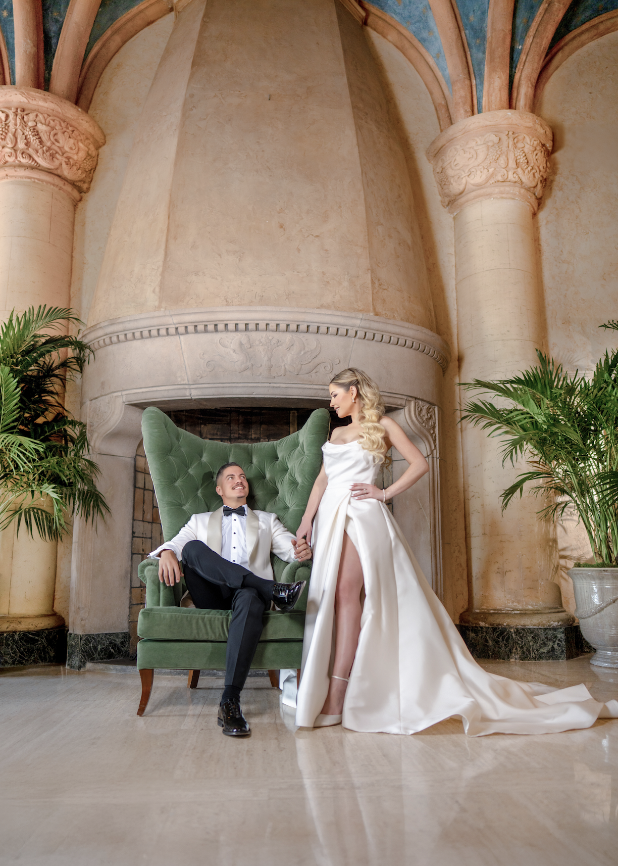 A bride and groom in wedding attire pose in front of a fireplace with large columns and plants on either side.