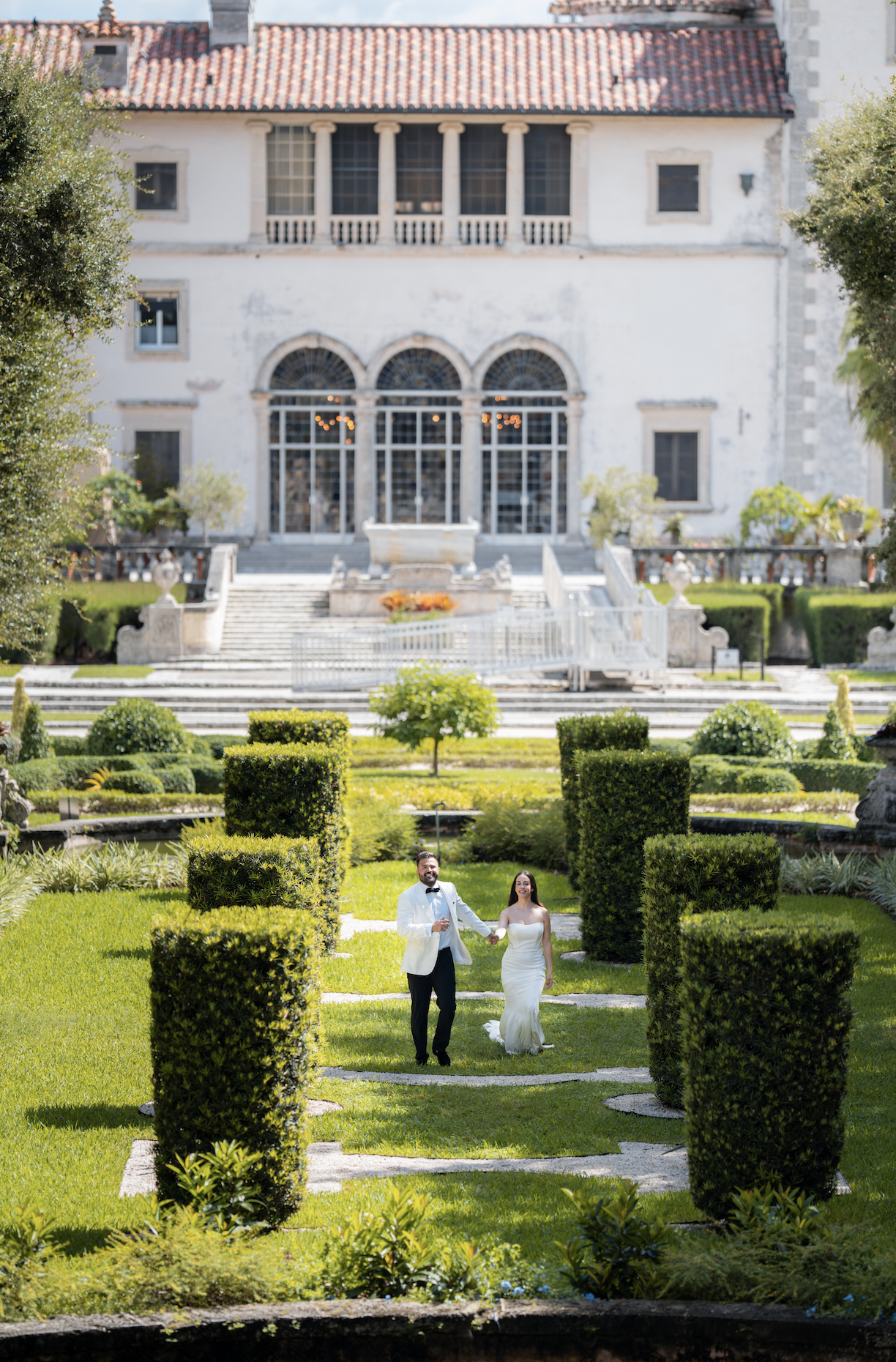 A couple in wedding attire walking through a lush, manicured garden with neatly trimmed bushes and trees, in front of an elegant, historic building with large arched windows and a staircase.