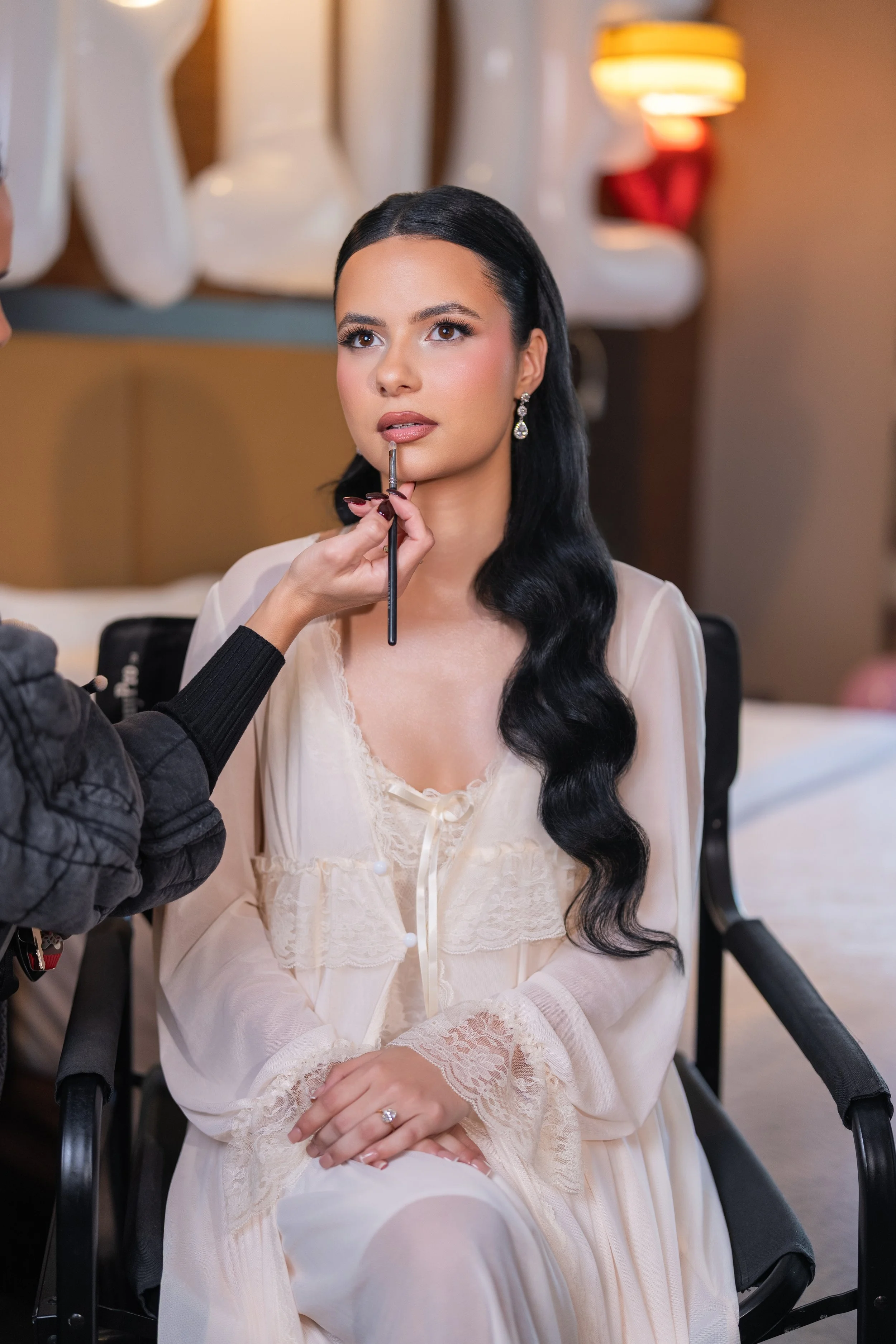 A woman sitting down getting her makeup done with a makeup brush near her lips, wearing an elegant cream-colored lace robe, and sitting in a room with warm lighting and decorative elements.