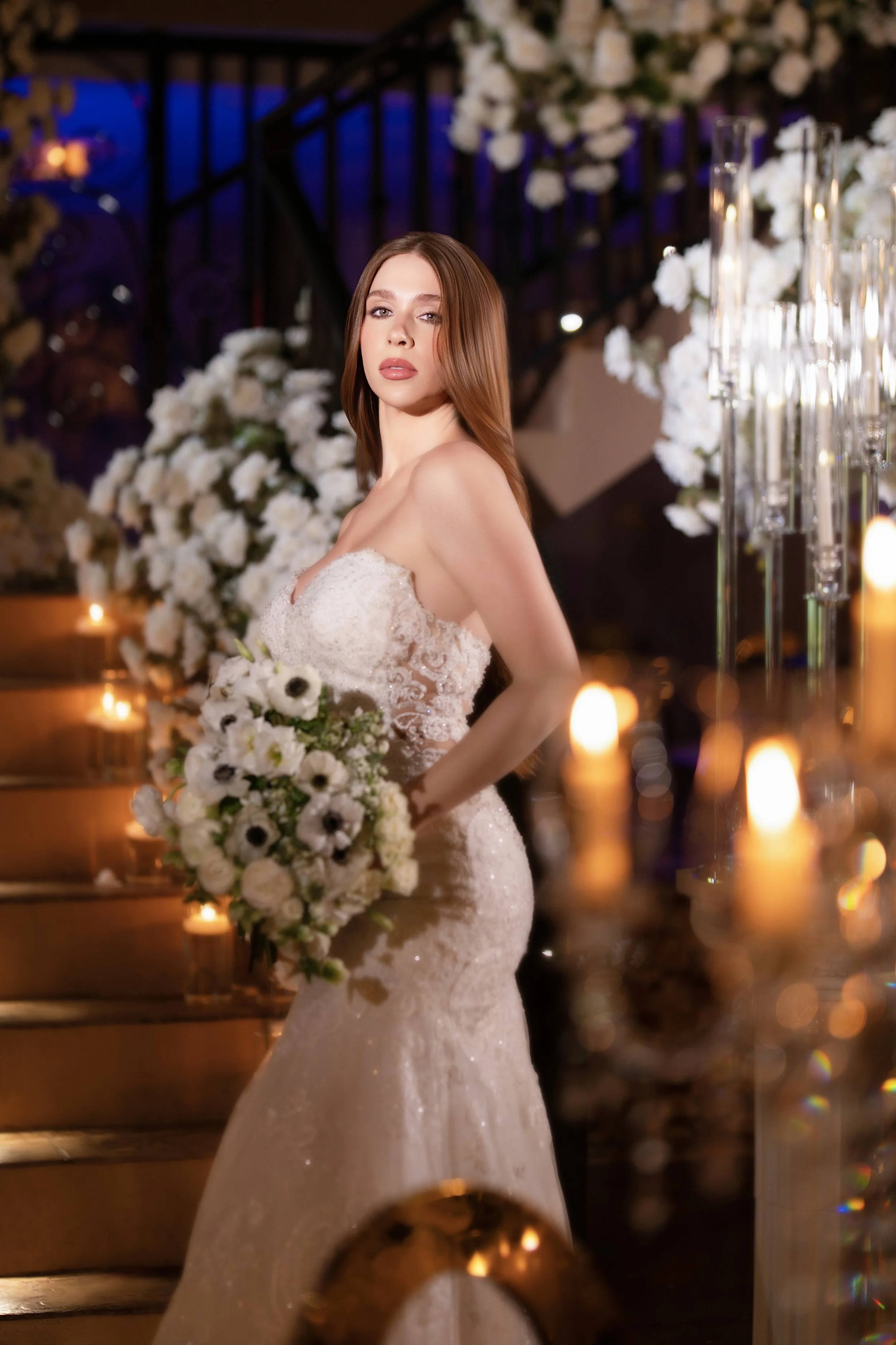 A bride in a strapless wedding gown holding a bouquet of white flowers, standing in a decorated venue with candles and white floral arrangements.