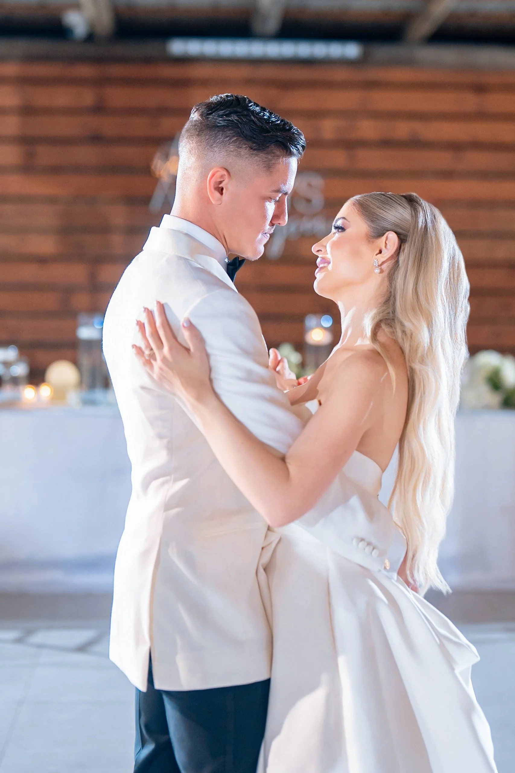 A couple dancing at a wedding reception, gazing into each other's eyes, with wooden walls and candlelit tables in the background.