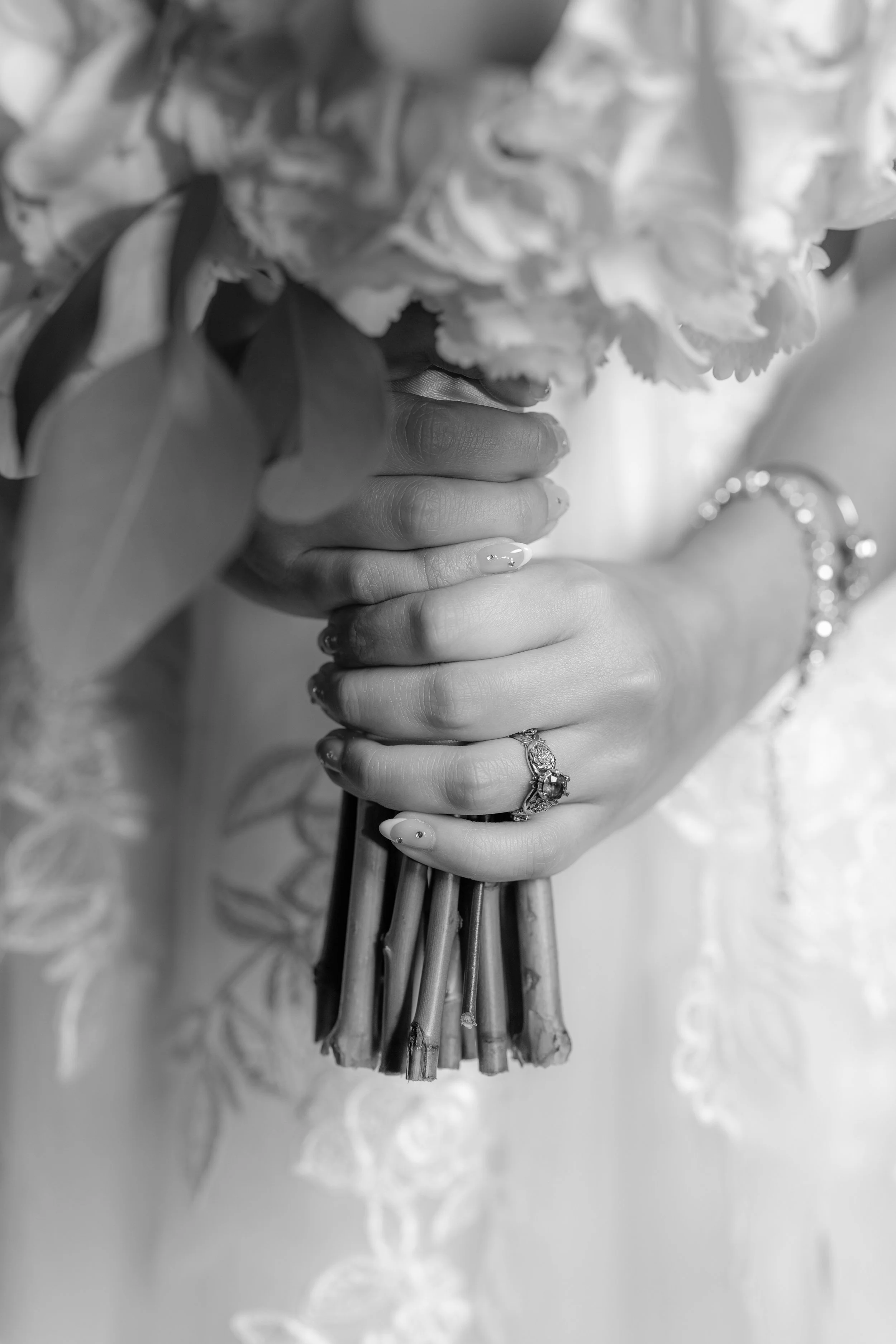 Close-up of a woman's hand holding a bouquet of flowers, showing rings and bracelets, with lace fabric in the background.