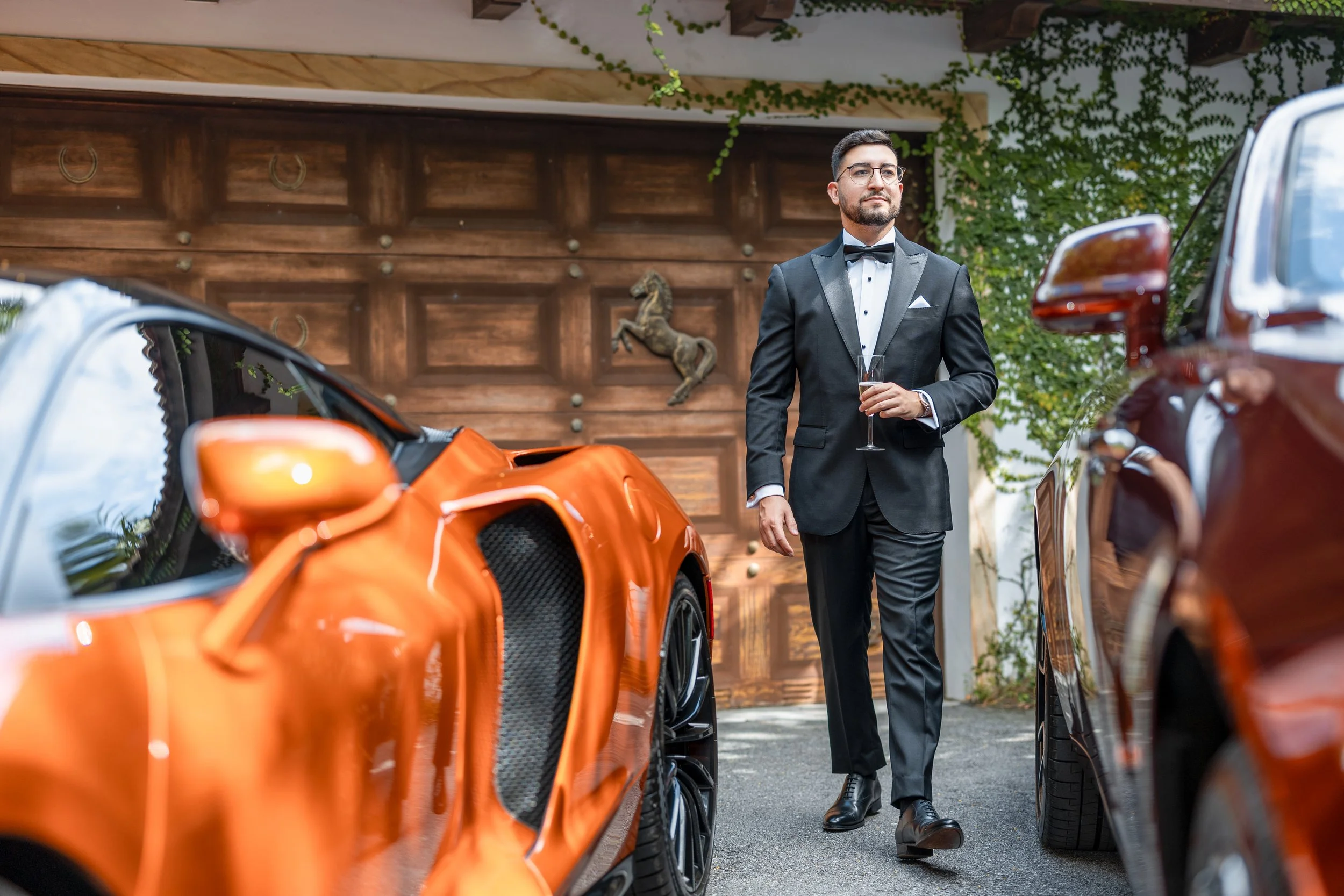 A man in a black tuxedo with a bow tie and glasses walking between two luxury cars, holding a glass of champagne, in front of a wooden garage door with a horse decorative piece.