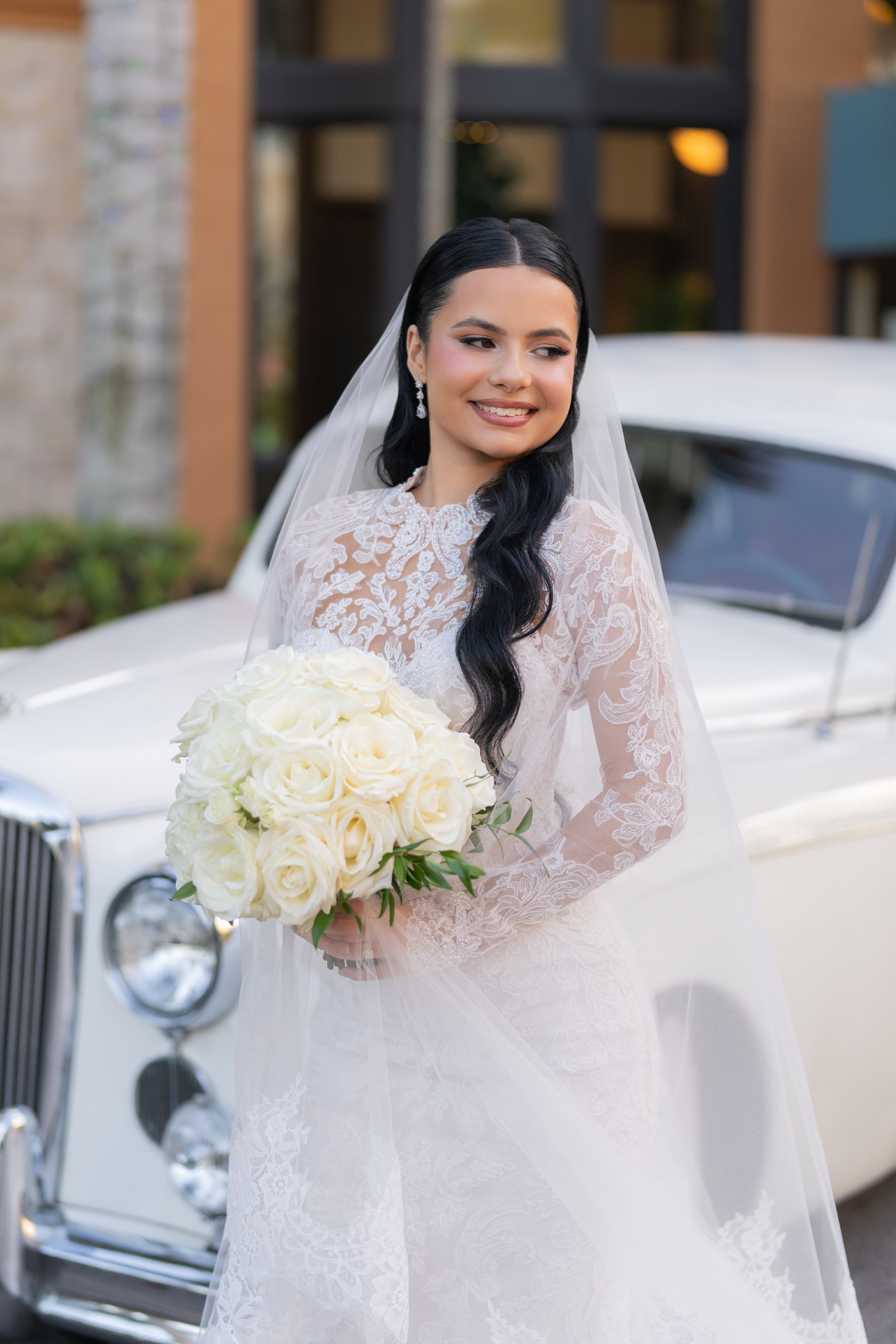 A bride in a white lace wedding dress holding a bouquet of white roses, standing in front of a classic white car.