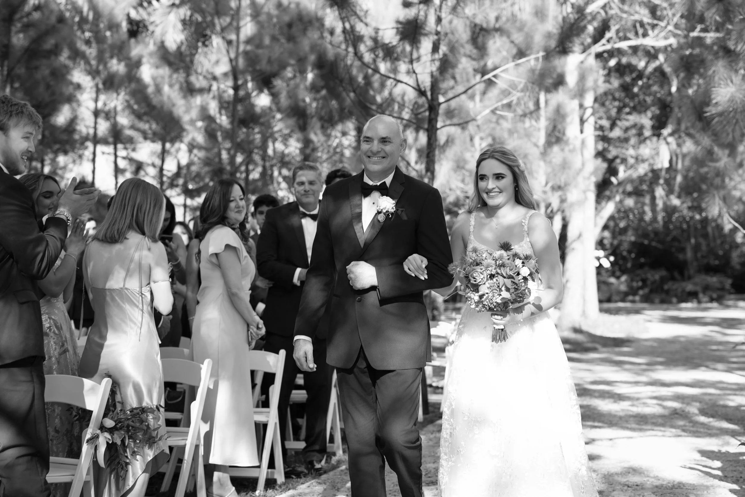 A bride and groom walking arm-in-arm outdoors during their wedding ceremony, with guests clapping and smiling in the background, surrounded by trees.