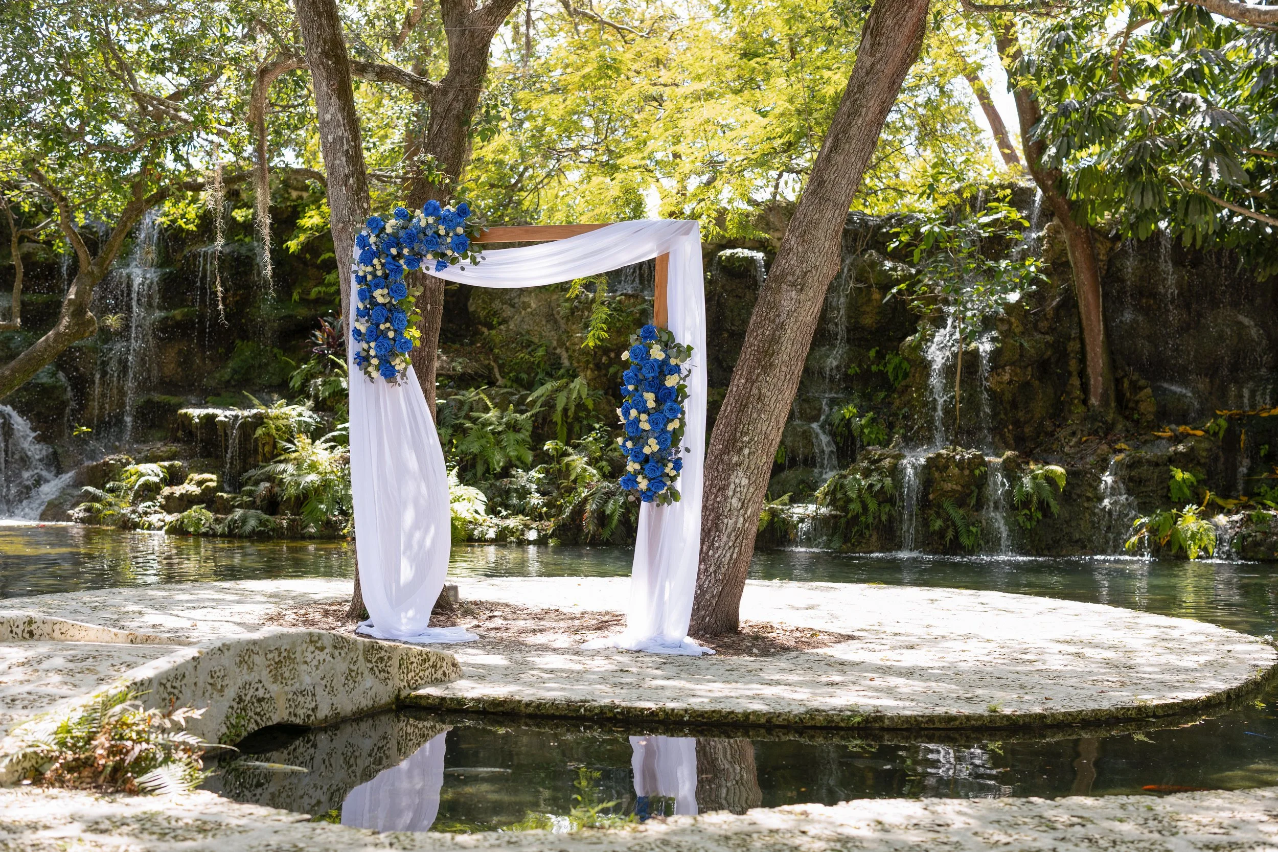 A wedding arch decorated with white fabric and blue flowers, set in a lush outdoor area with trees, waterfalls, and a small pond.