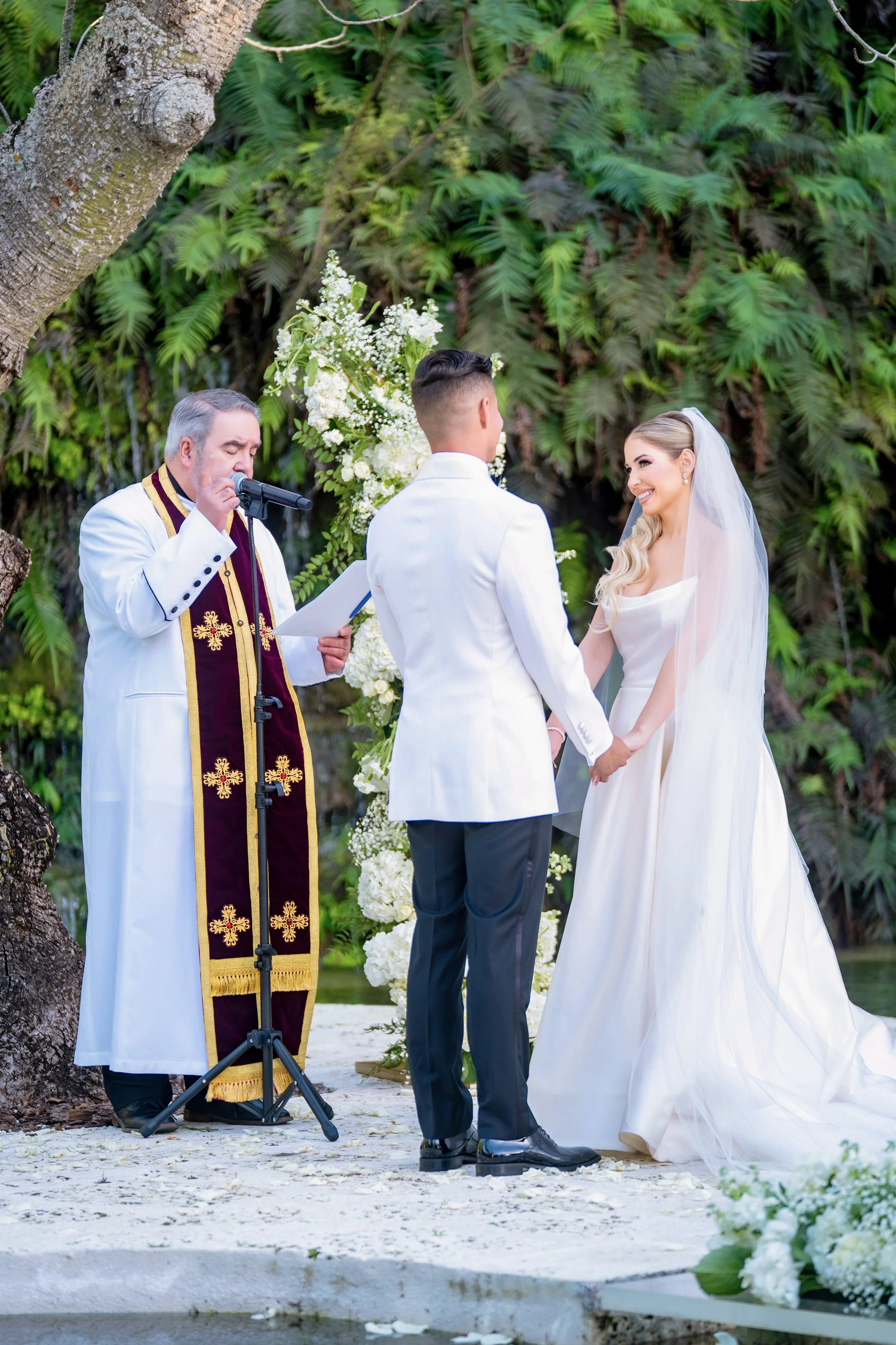 A couple getting married outdoors, holding hands and facing each other, with an officiant reading from a paper. The scene is decorated with white flowers and surrounded by lush greenery.