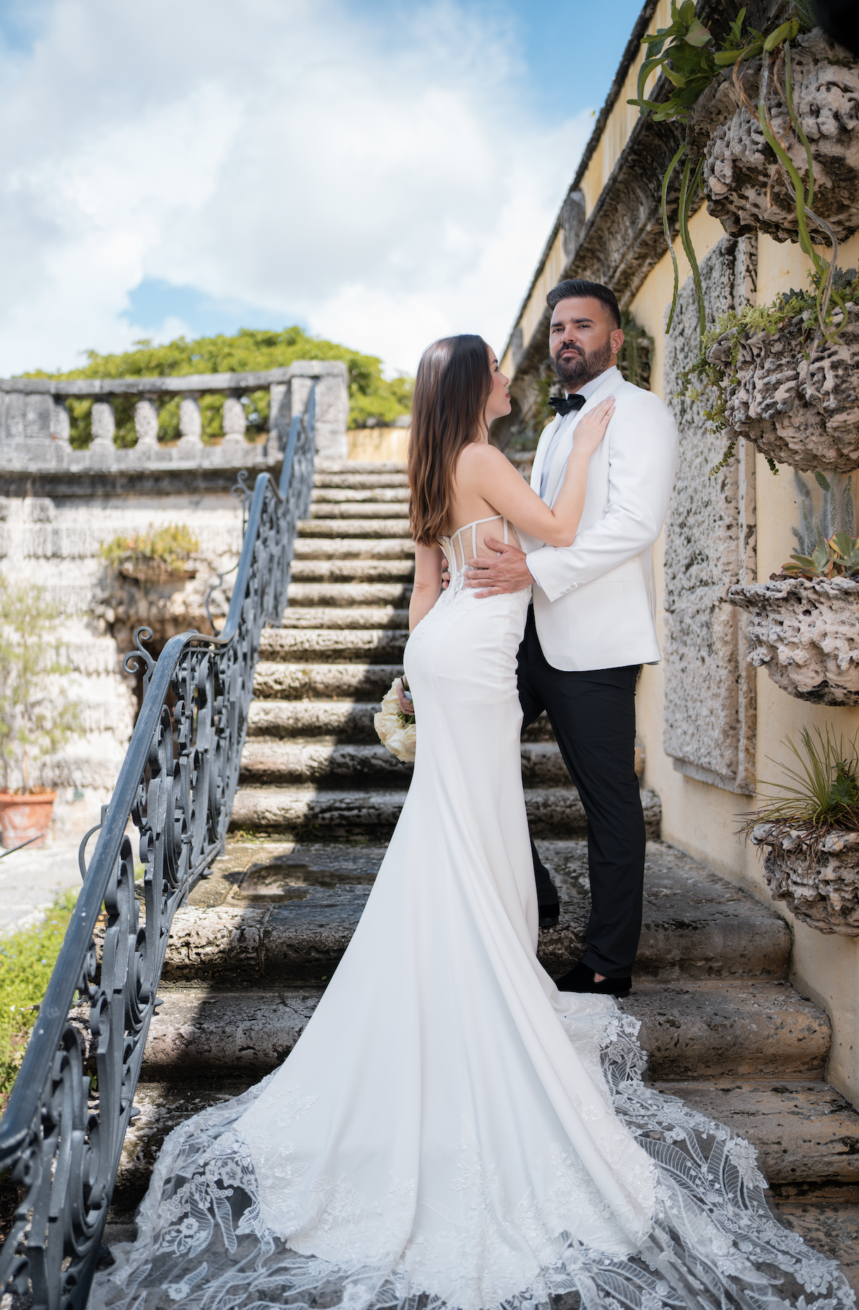 A couple dressed in formal wedding attire, with the woman in a white wedding gown and the man in a white tuxedo jacket, pose on stone stairs outdoors, surrounded by greenery and a stone wall.