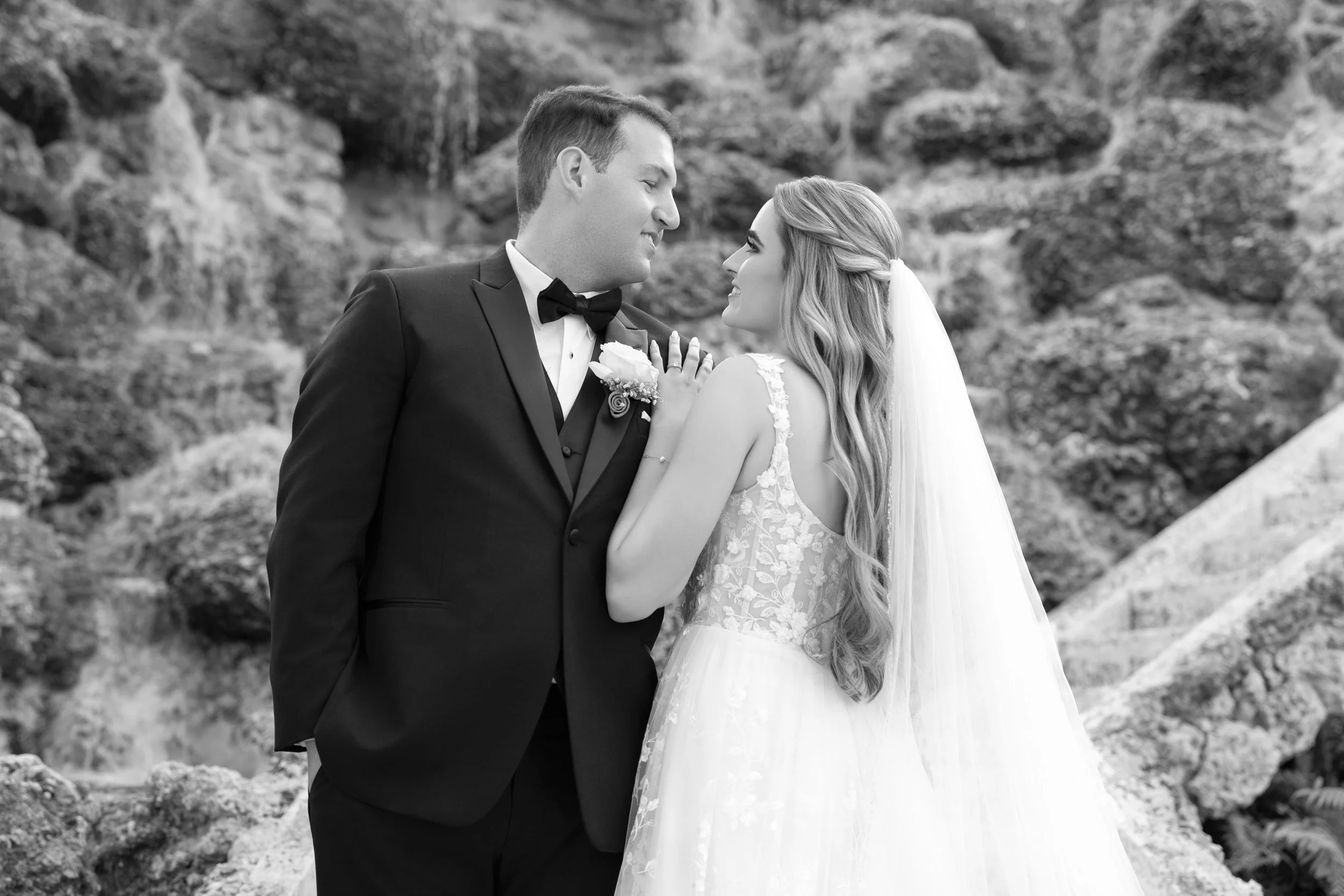 A black and white photo of a bride and groom standing close together outdoors, gazing into each other's eyes. The groom wears a tuxedo with a bow tie, while the bride wears a lace wedding gown with a veil and has her hand on his chest. They are surro