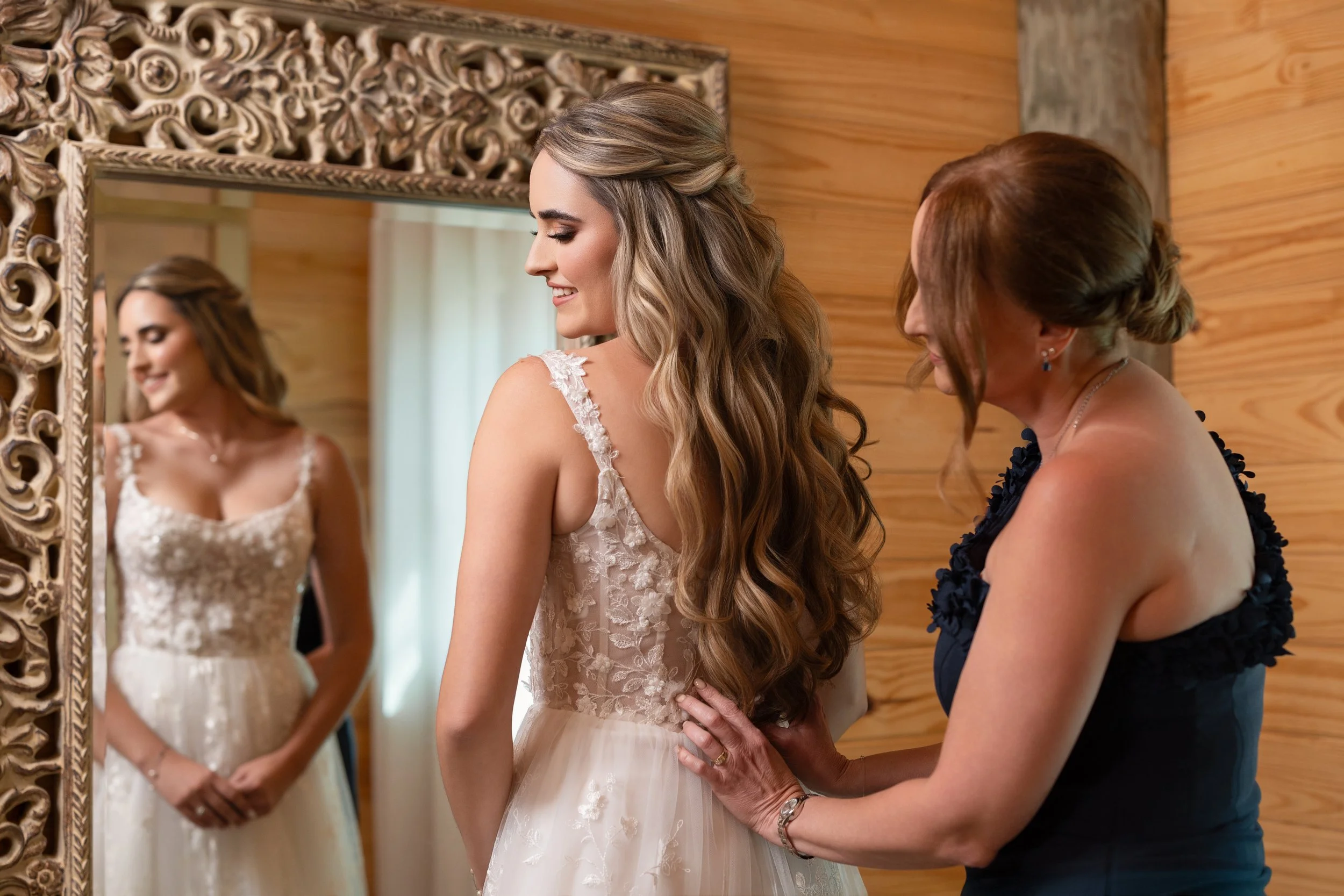 A bride and her mother preparing for her wedding in a cozy, wooden room. The bride is wearing a white wedding dress with lace on the top, and the mother is in a black dress with floral details, crouching to adjust the bride's dress while they look at
