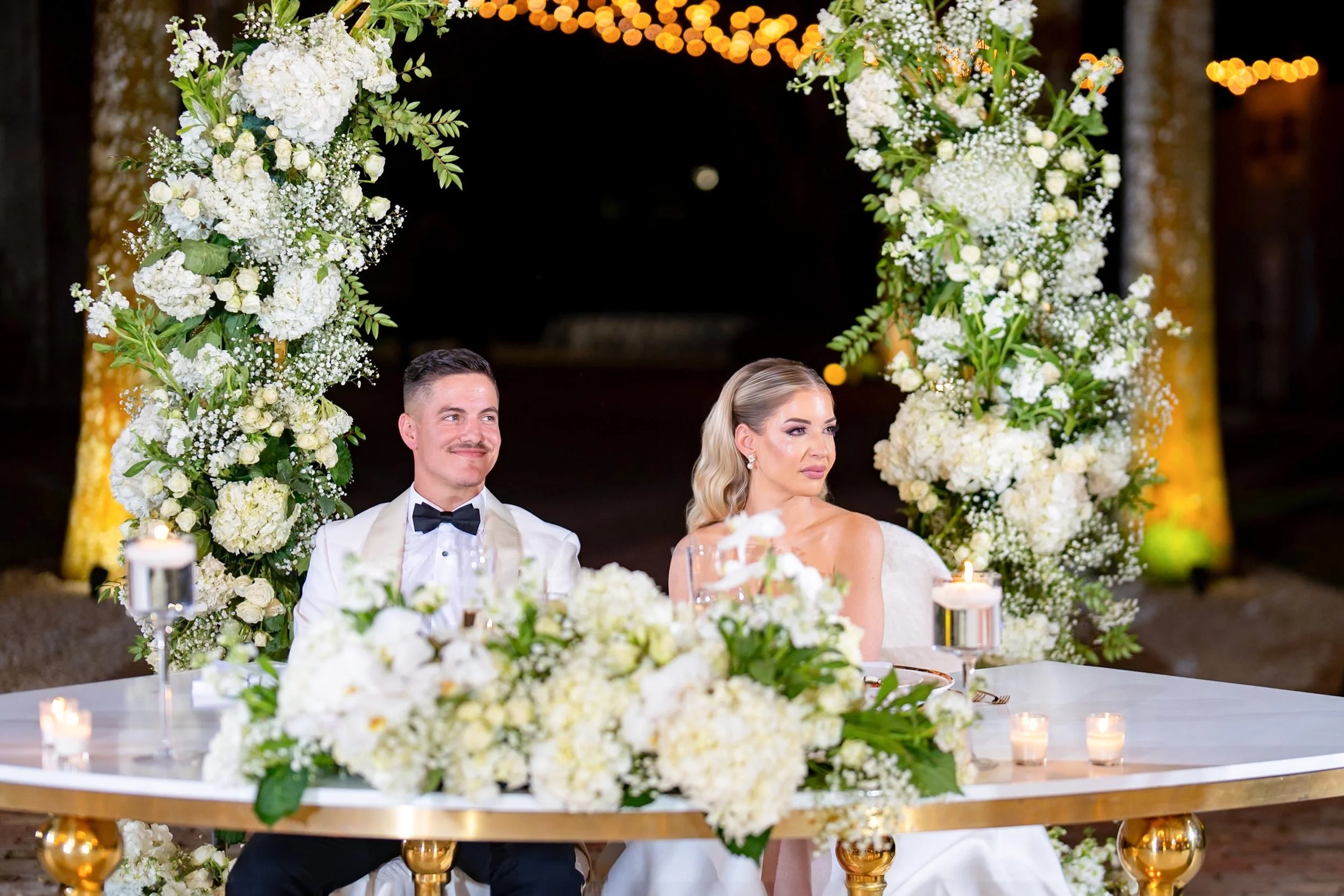 A bride and groom sitting at a wedding reception table decorated with white flowers and candles, in front of a floral arch at night.