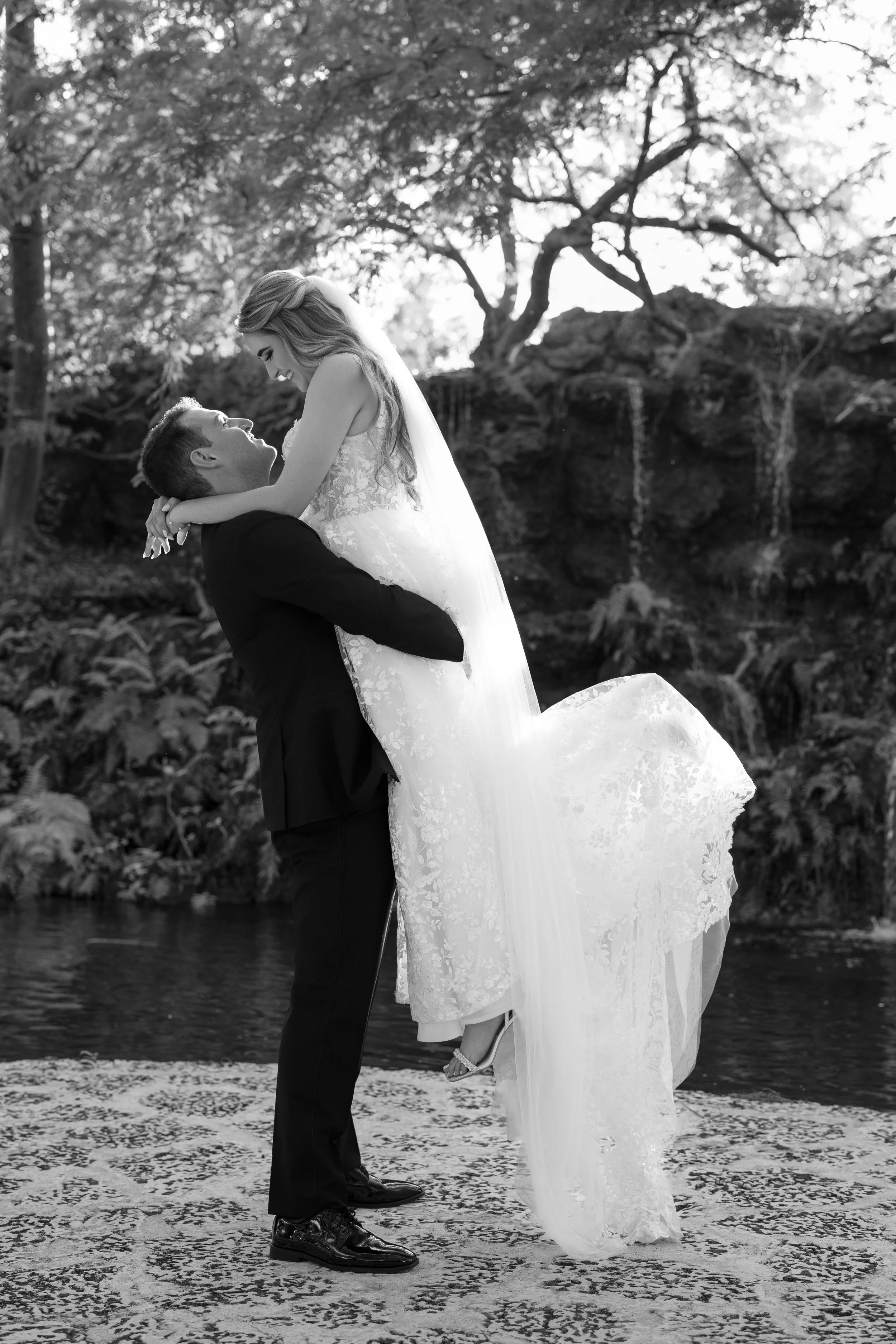 A Black and white photograph of a wedding couple, with the man lifting the woman in his arms near a pond or stream, surrounded by trees and natural scenery.