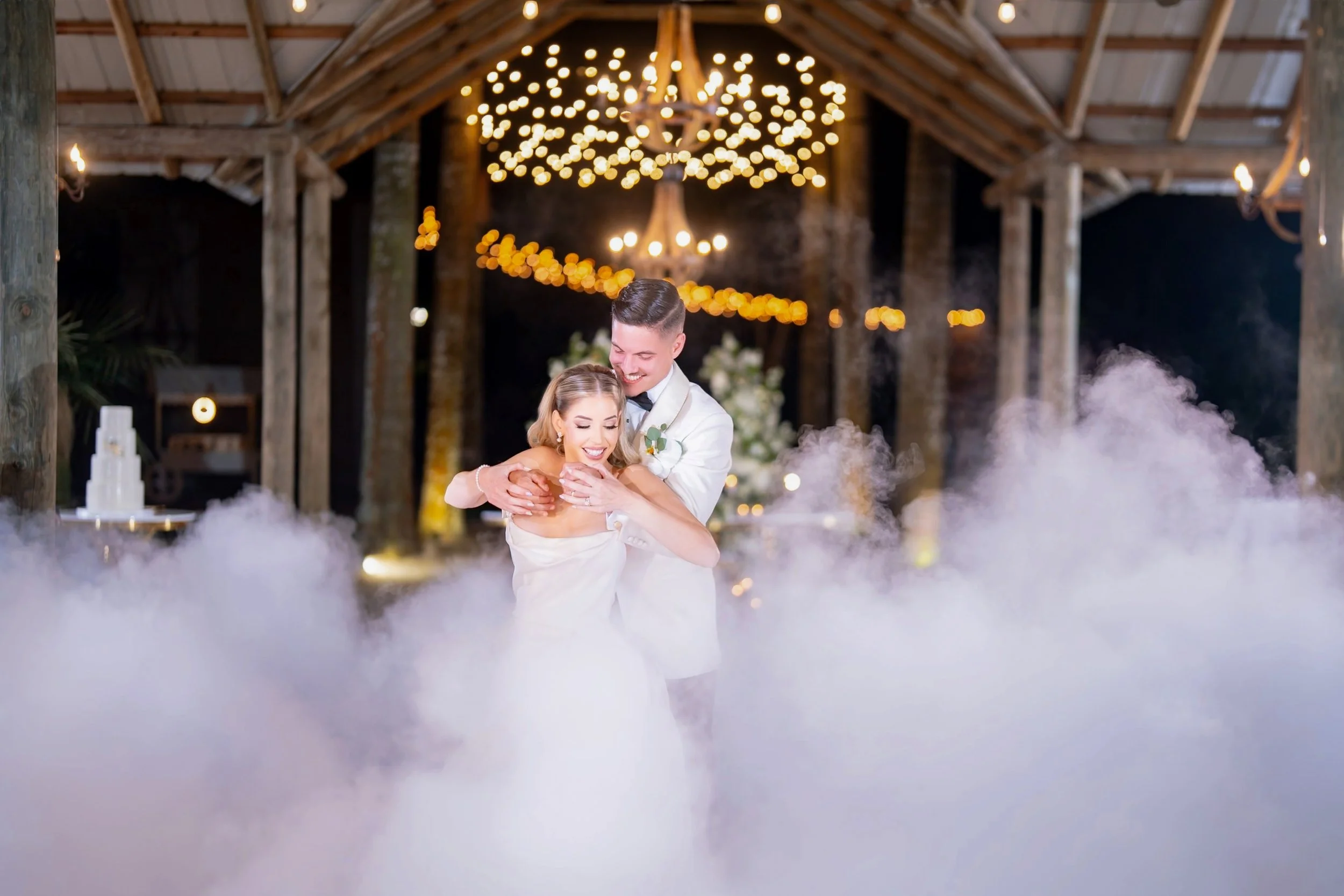 A newlywed couple dancing at their wedding reception in a rustic barn decorated with string lights, surrounded by fog.