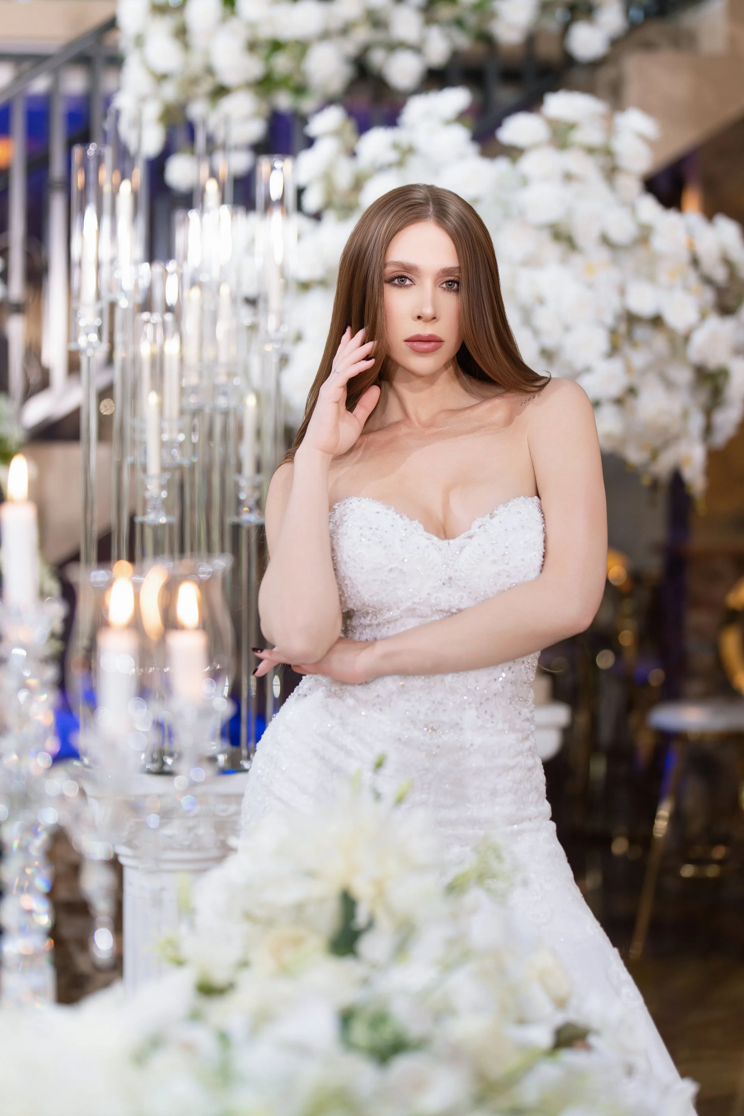 A woman in a white wedding dress standing indoors with white flowers and candles around her.