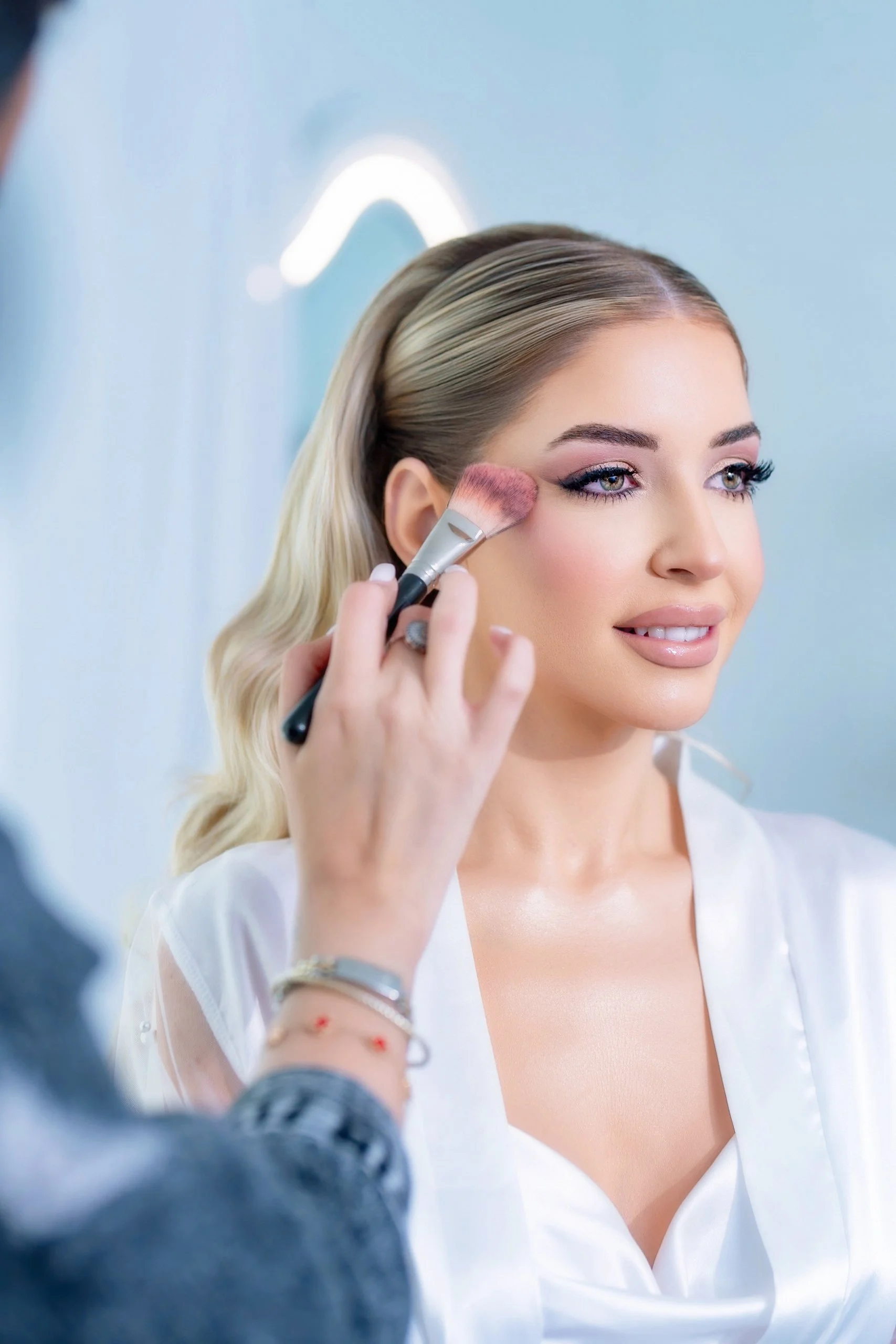 A woman is having makeup applied to her face, with a focus on her eye area, by a makeup artist using a pink makeup brush.