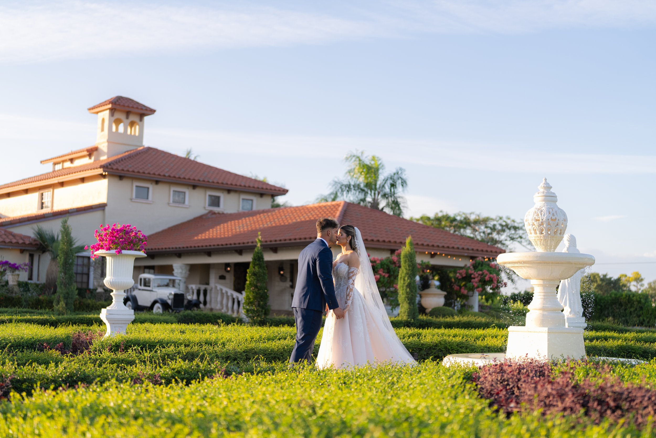 A bride and groom in wedding attire holding hands and touching foreheads in a garden with decorative urns and a large building with red-tiled roof in the background.