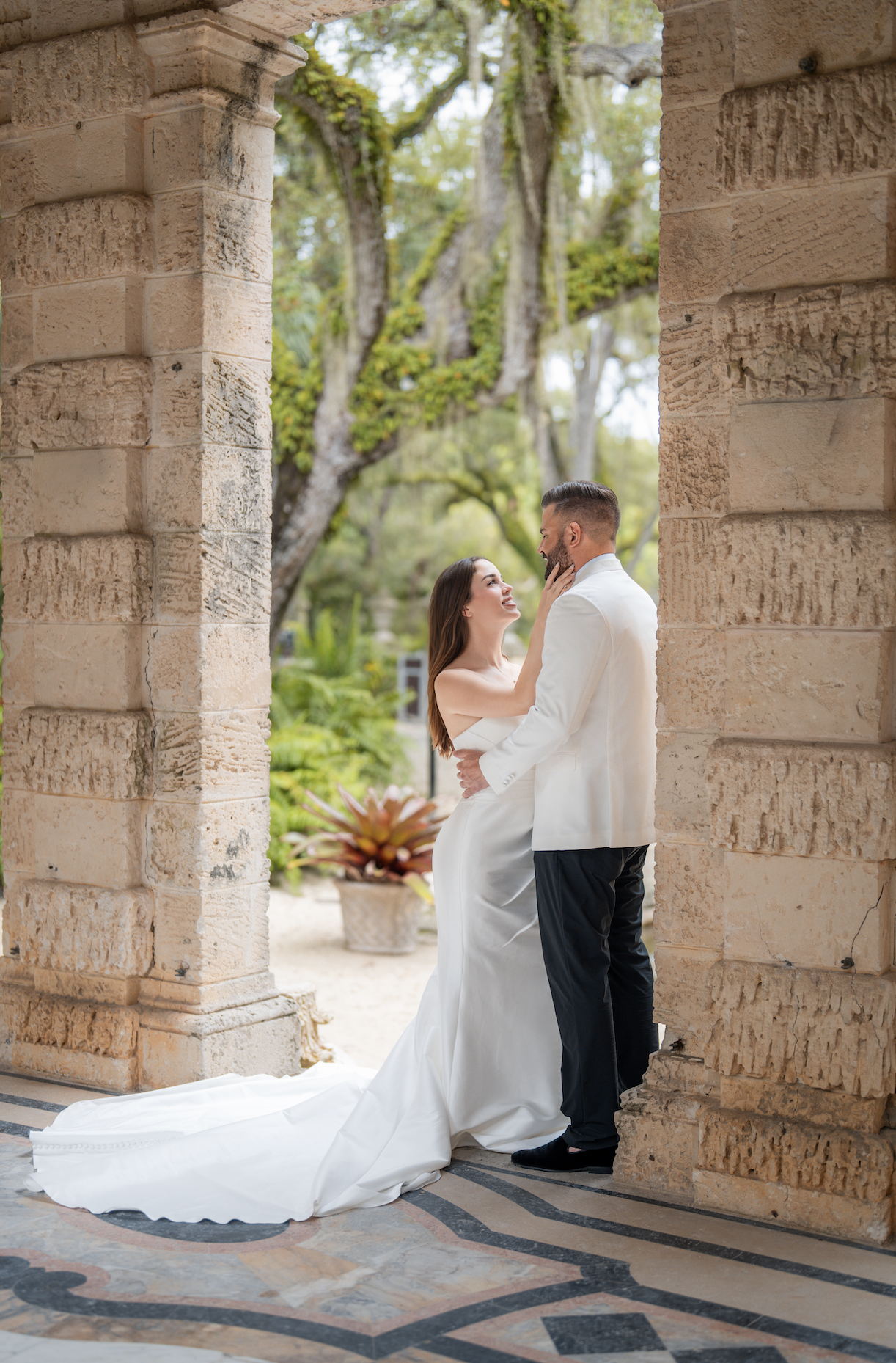 A bride and groom share a romantic moment under stone archways on their wedding day, with greenery and trees in the background.